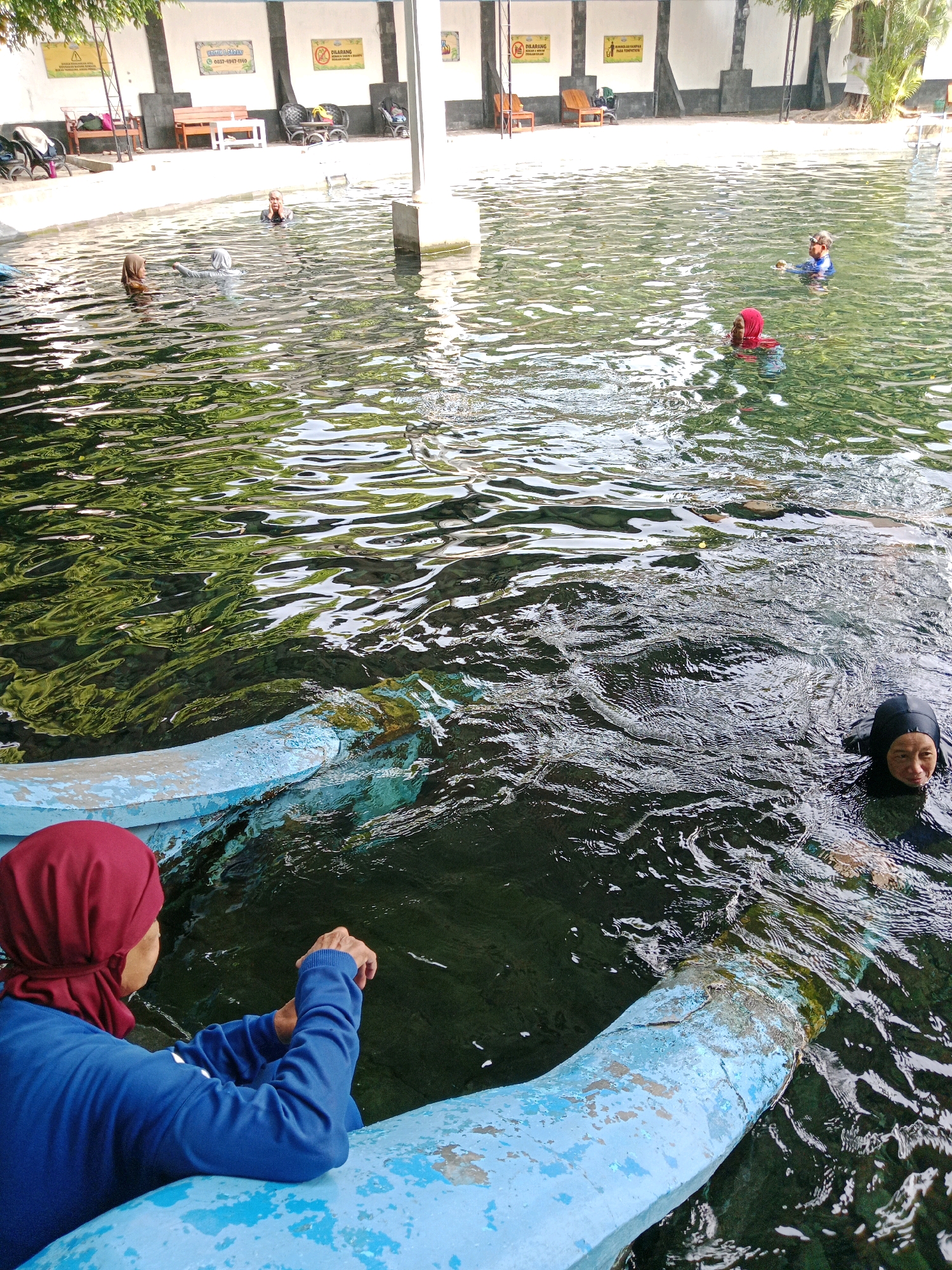 The image depicts an outdoor swimming pool facility in Jentekan, Indonesia, with several individuals present in the water. In the foreground, a person wearing a red hijab and a blue long-sleeved shirt is leaning on the edge of the pool, facing away from the camera and looking into the water. To the right, another person in a black swimming cap is partially submerged in the water, looking towards the camera with a smile. In the middle ground, two individuals are visible in the water, one wearing a light-colored hijab and the other a darker one, appearing to be conversing. Further in the background, a person in a blue swimming suit is swimming. The pool is surrounded by a concrete walkway and a wall with signage. Trees and shaded areas are visible in the background, suggesting a recreational setting. The water is clear and appears to be reflecting the surrounding greenery and structures.