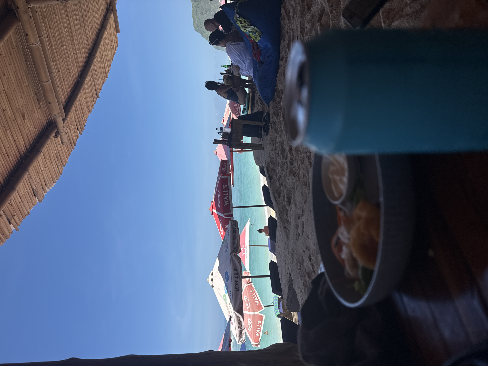 The scene depicts a beachfront eatery in Kuta Selatan, Indonesia. The view, framed by a bamboo roof and obscured by a foregrounded blue water bottle and a plate of food, reveals beachgoers relaxing on the sand. Advertisements for "WALL'S" and "BINTANG" are prominently featured on the beach umbrellas. People are sitting at tables near the beach, some using bean bags. The ocean stretches out beyond the beach, and hills can be seen in the distance.