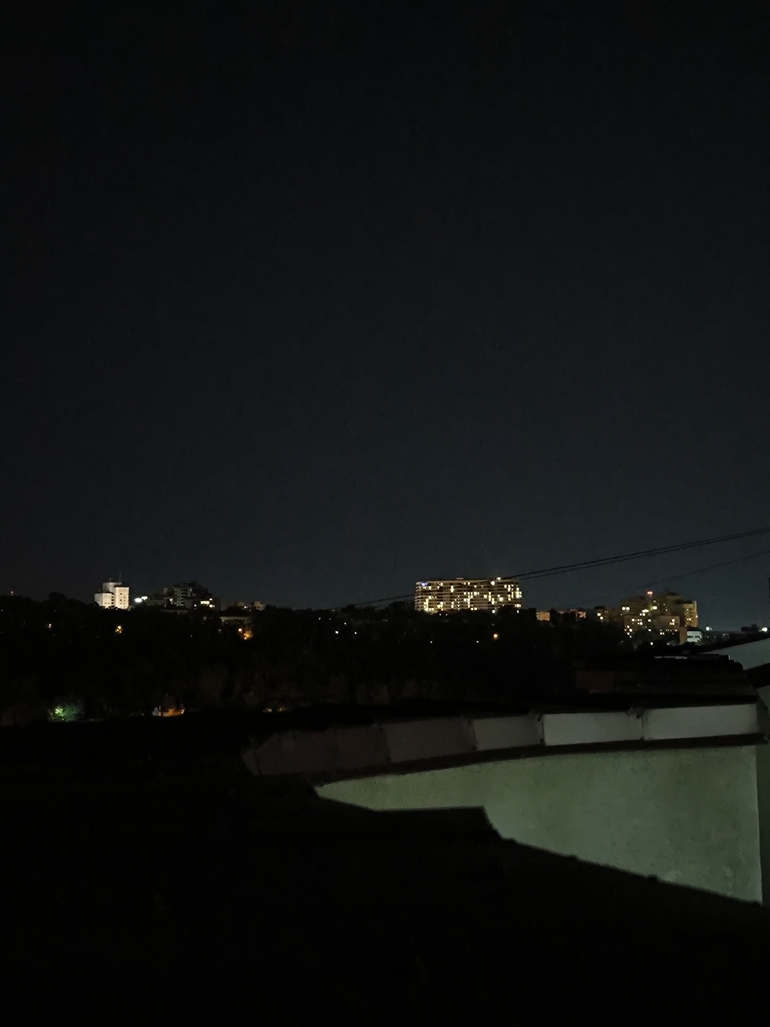 The image shows a nighttime cityscape. In the distance, several buildings are illuminated by artificial lights, suggesting an urban environment. The foreground is dominated by the dark silhouettes of rooftops and structures. The sky is dark and clear, indicating a lack of clouds and minimal light pollution. The scene appears to be from a high vantage point, looking out over the city. Without further context or identifying landmarks, it is impossible to definitively pinpoint the specific location within Chișinău, Moldova.