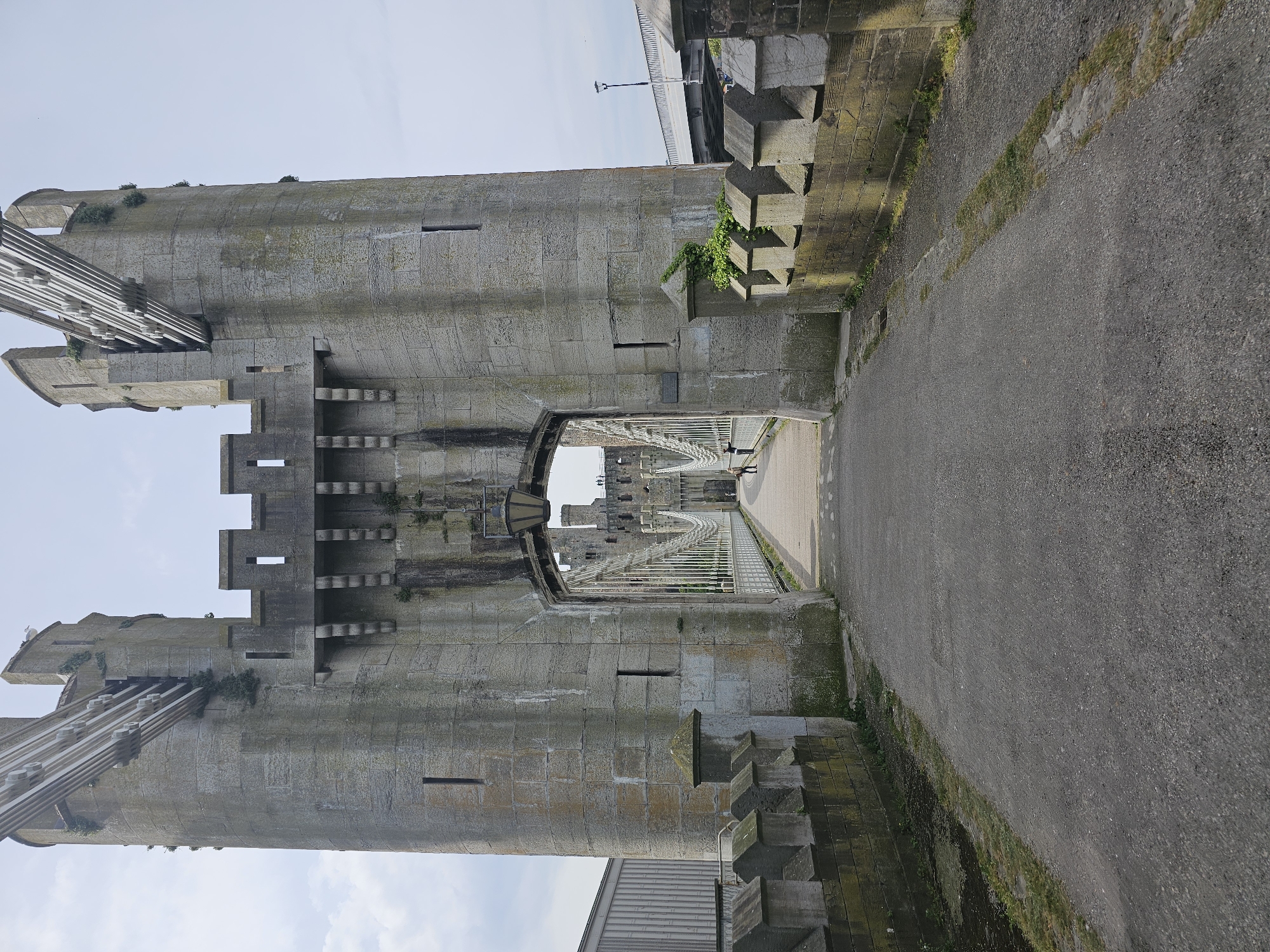 The image captures a view of Conwy Castle in Llandudno Junction, United Kingdom. The scene is viewed through a stone archway of the castle. The archway leads to a paved walkway and a bridge stretching into the distance, passing through additional arches of the castle. The castle walls are constructed from large grey stones, showing signs of age and weathering, with green moss growing in places. There are crenellations on the towers. The paved walkway is made of a grey material, possibly asphalt or concrete, with patches of grass along the edges. A few people are walking on the bridge in the distance. A modern bridge is seen overhead, partially obscured by the castle walls. The sky above is pale blue with a few clouds.