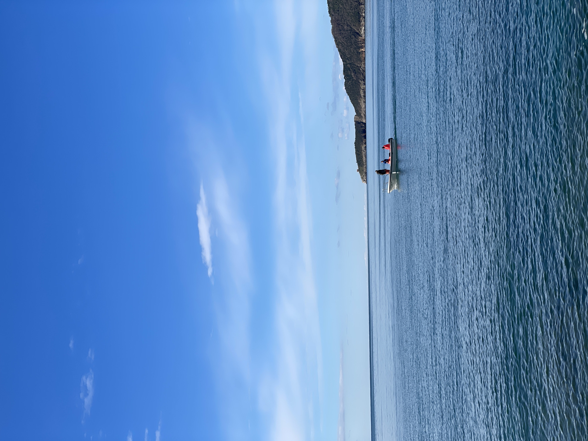 Three people are in a white rowboat on calm, blue water. A cliff face with sparse vegetation is visible on the horizon. The sky is blue with thin white clouds. The location appears to be Rafailovići, Montenegro, given the coastal landscape.