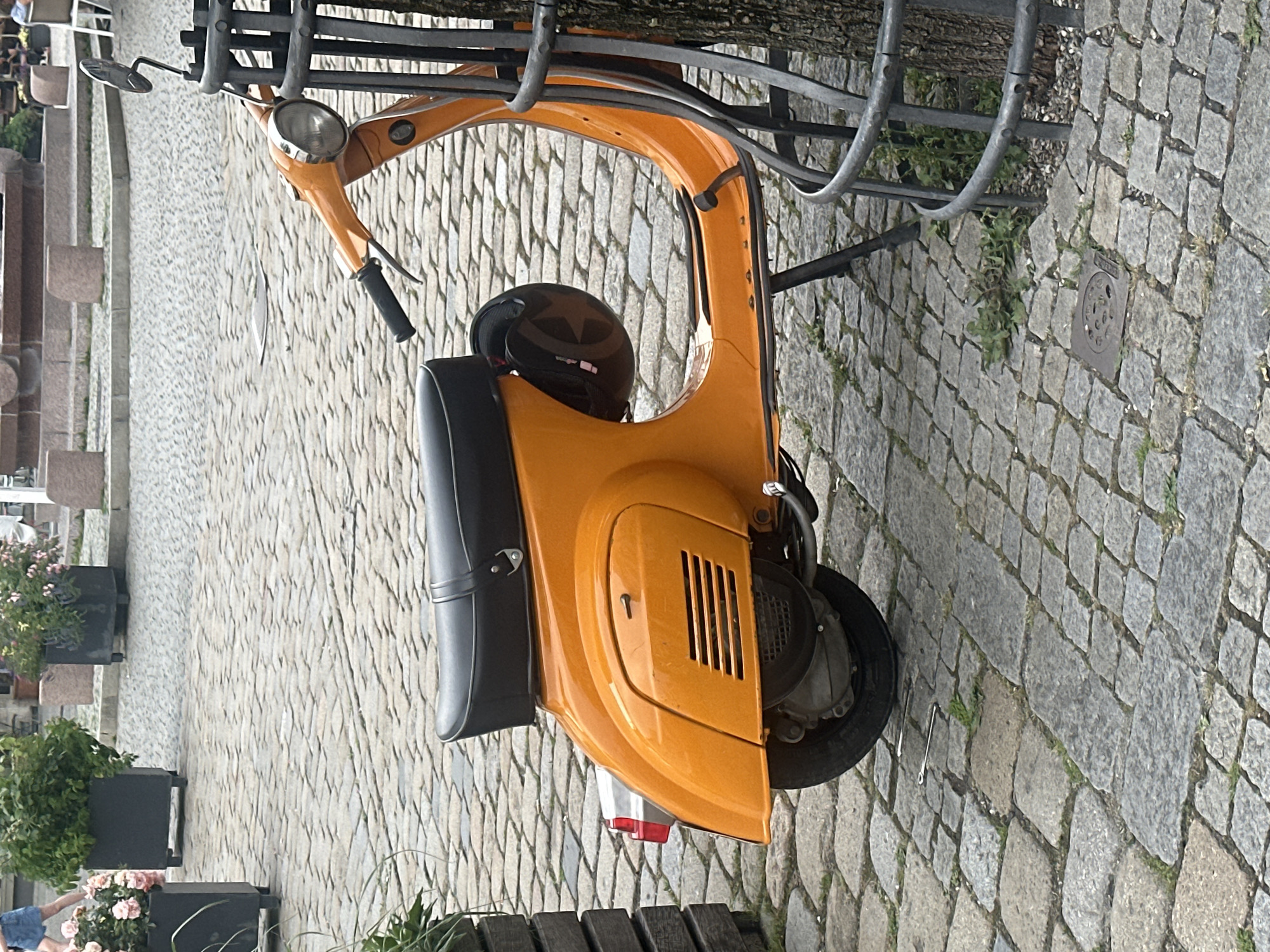An orange Vespa scooter is parked on a cobblestone street in an urban environment. The scooter is facing right and is partially obscured by a metal railing and a tree. A black helmet is hanging from the back of the scooter. In the background, there are planters with greenery and flowers, and a fountain structure. The ground is paved with grey cobblestones. The scene is outdoors, possibly in a public square. There are no people clearly visible in the image.