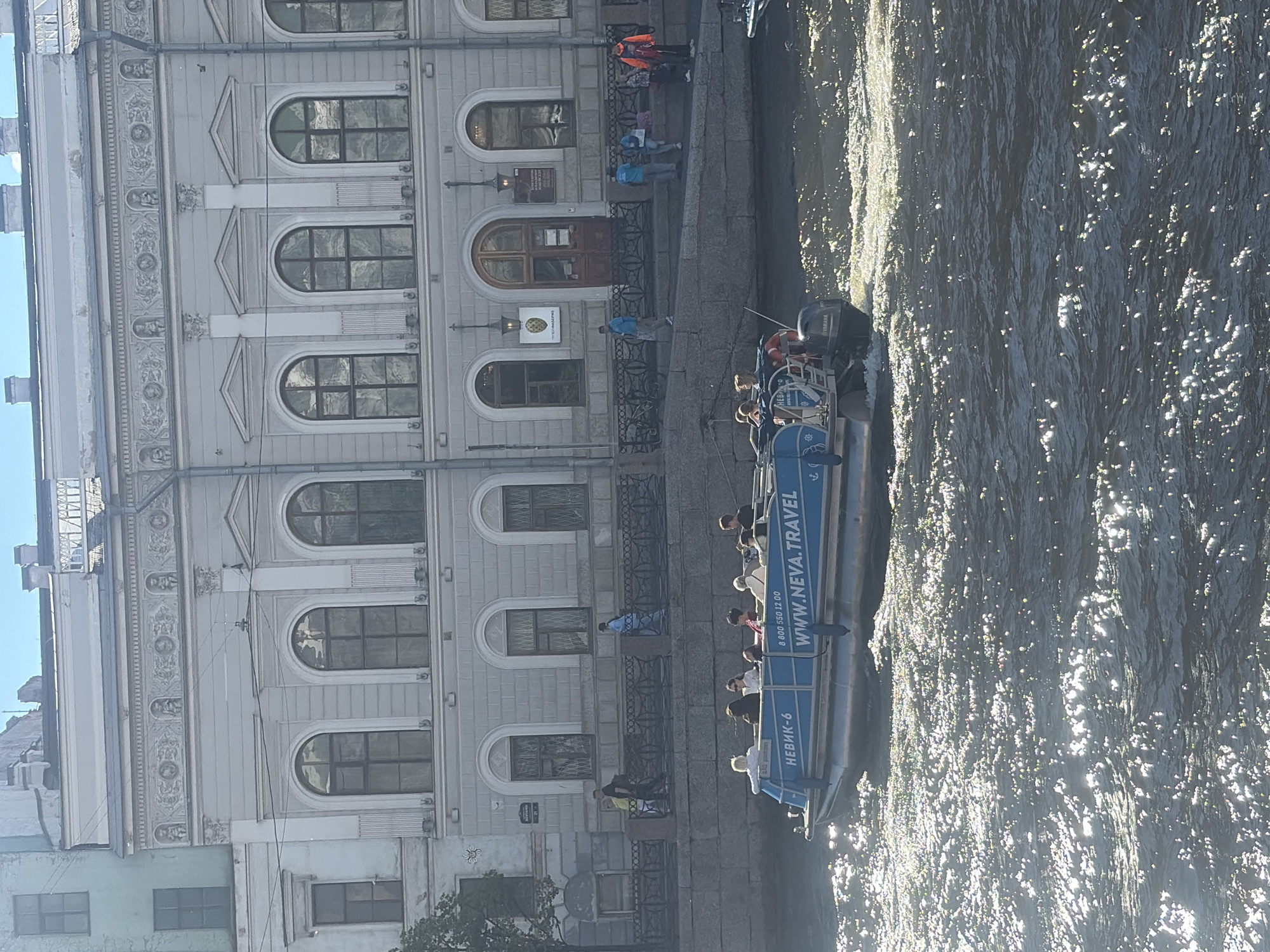 This is a photograph taken from a boat on a canal in Saint Petersburg, Russia. The boat is a tour boat with several passengers. The building beside the canal is an old, ornate building with arched windows. The water in the canal is sparkling with sunlight, suggesting it is daytime and sunny. The overall mood is one of leisure and sightseeing.
On the side of the boat, the text "WWW.NEVA.TRAVEL" is visible, along with a phone number "8 800 530 12 00" and the word "НЕВИК-6". The people on the boat appear to be tourists enjoying the scenery. Some are standing and looking around, while others are seated. The building displays classical architecture with decorative elements and many windows. The canal's surface reflects the sunlight, creating a dappled effect. The scene captures a typical tourist experience in the historic city of Saint Petersburg.