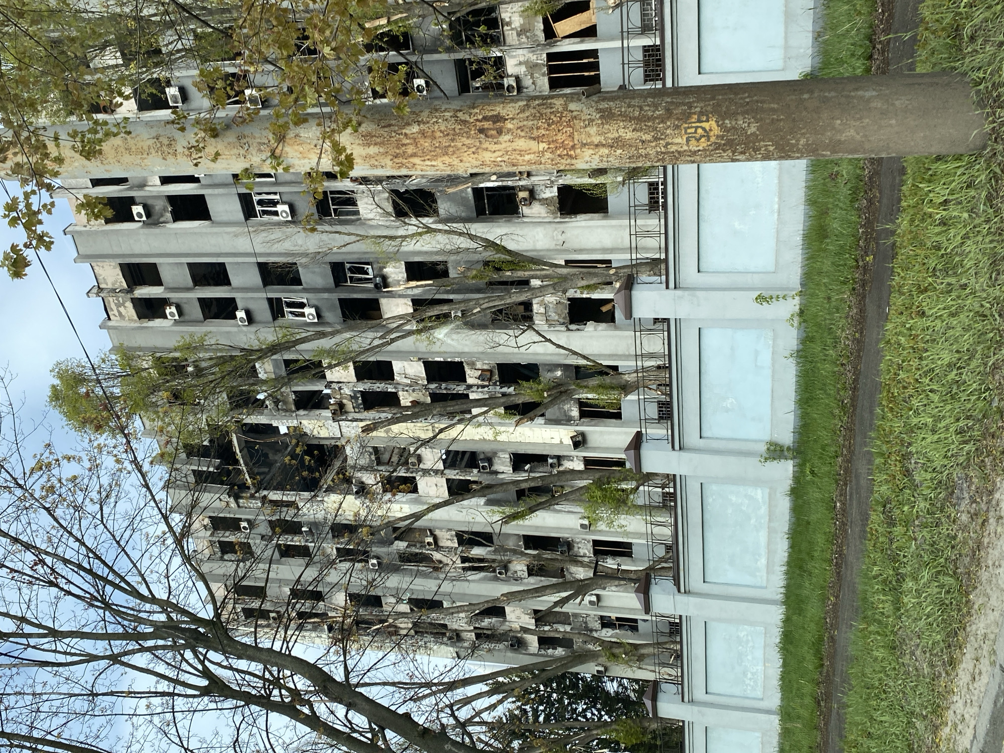 The image shows a tall, severely damaged building, likely an apartment complex, in an urban environment. The building exhibits extensive fire damage, with blackened window openings and scorched facades. Many windows are missing entirely, while air conditioning units are visible on the exterior walls. A concrete barrier or wall runs along the base of the building, paralleled by a narrow sidewalk and grassy area. In the foreground, bare tree branches partially obscure the view of the building. A utility pole stands to the right of the building. The location is Unknown, Unknown.