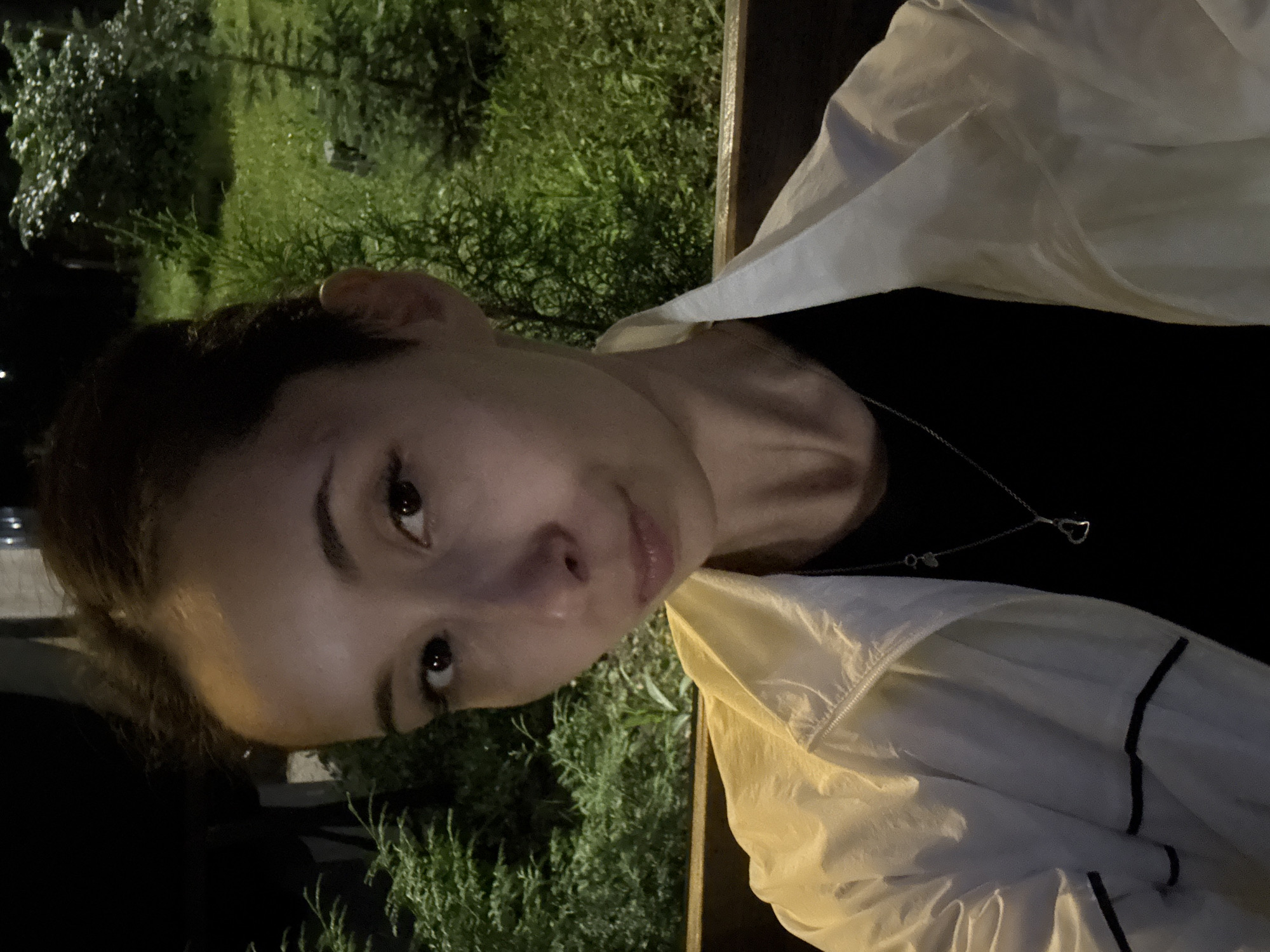 A close-up, waist-up, selfie-style photograph features a young woman sitting on a wooden bench outdoors at night. She has dark hair pulled back and is looking directly at the camera with a neutral expression. She is wearing a black t-shirt and a light-colored jacket with black trim. A silver necklace with a heart pendant is visible. The background consists of dark foliage and grass, with some artificial lighting illuminating the scene, suggesting an urban park or garden setting. The location is identified as Doma 1096 Km, Russia.