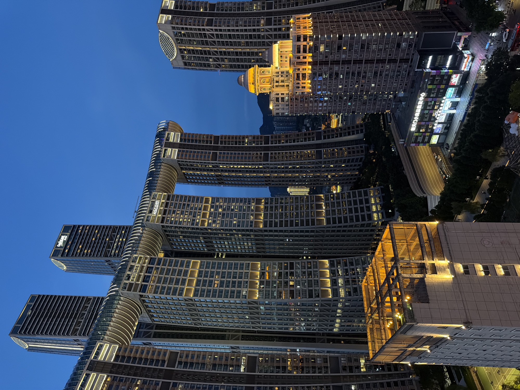 This is a high-angle, dusk photograph of the Raffles City Chongqing complex, located in the Yuzhong District of Chongqing, China. The architectural complex is dominated by its distinctive interconnected skyscrapers, featuring a horizontal structure resembling a ship or a dragon, known as the "Crystal". The exterior of the structures is illuminated with warm, golden lights, highlighting the modern architectural design. In the foreground, a lighter-colored building with a grid-like roof structure is also lit from within. Below the main complex, other buildings of varying heights and designs are visible, some displaying colorful signage. The sky is a deep blue, indicating the transition from day to night. Shadows are cast by the buildings, and the overall scene depicts an urban landscape at twilight, emphasizing the illuminated modern architecture. No individuals are clearly discernible in the image.