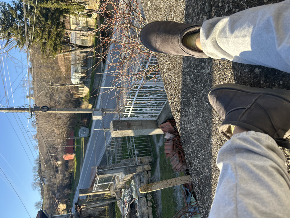 The image, captured from an elevated, first-person perspective, depicts the lower legs and feet of an individual resting on a textured, grey concrete surface. The person is attired in light grey sweatpants and dark brown Ugg-style boots, revealing yellow/lime green socks at the ankles.
Below the foreground concrete surface, a concrete pillar and white metal gate structure are visible, flanked by sections of a low stone wall topped with chain-link fencing. A pile of terracotta roof tiles rests on the ground directly beneath this fence line.
In the midground, a paved two-lane road traverses the scene horizontally. A grey metal dumpster is situated on the road's verge to the right. Multiple utility poles carrying power lines extend upwards from the roadside.
The background features a gently sloping hillside densely covered with bare deciduous trees, interspersed with some evergreen vegetation. Several residential structures with red-tiled roofs are visible among the trees, and a distinct red-colored building is located on the left slope of the hill. The sky is clear and blue, indicating daytime conditions with direct sunlight.
The depicted scene is located in Senovo, Bulgaria.