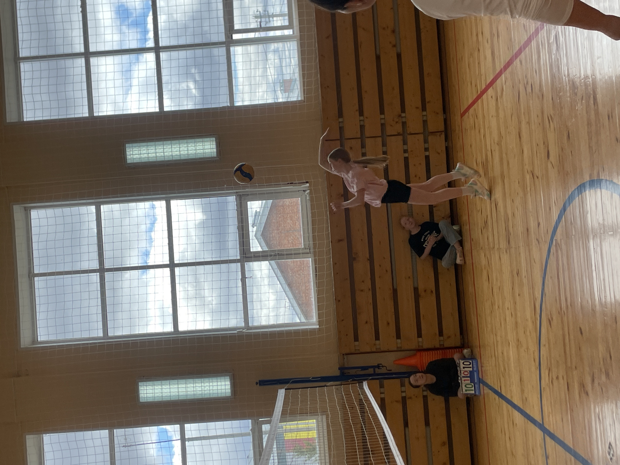 The image shows a volleyball game taking place indoors. A young girl in a pink shirt and black shorts is in mid-air, reaching to hit a volleyball. The game is happening in a gymnasium, as evidenced by the wooden floors, high ceilings, large windows with netting, and a volleyball net. A person stands near an orange cone holding a scoreboard with the numbers "10" visible. Another person is sitting on the floor. The windows reveal a cloudy sky, with brick buildings visible. There is no indication it is Подшивалово, Russia.