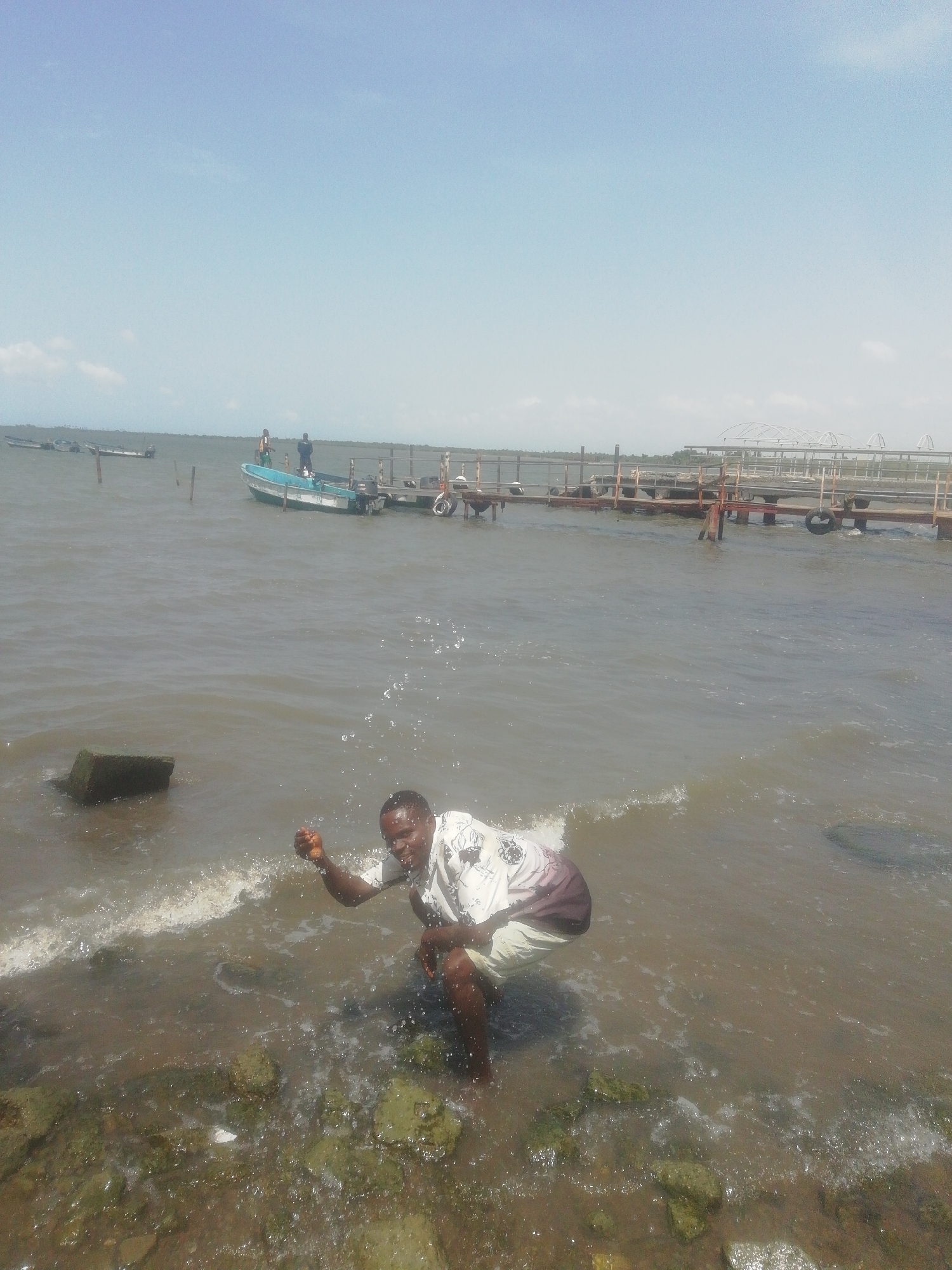 A man is squatting in the water along the shoreline of Ikorodu, Nigeria. He is wearing a short-sleeved, patterned shirt and shorts. The water is splashing around him, and he is holding his hand up as if he has just thrown something. Behind him, there is a pier extending into the water with boats docked alongside. Other boats are visible in the distance, and a few people are standing on the pier. The sky is clear and blue. Rocks and vegetation line the shoreline in the foreground.