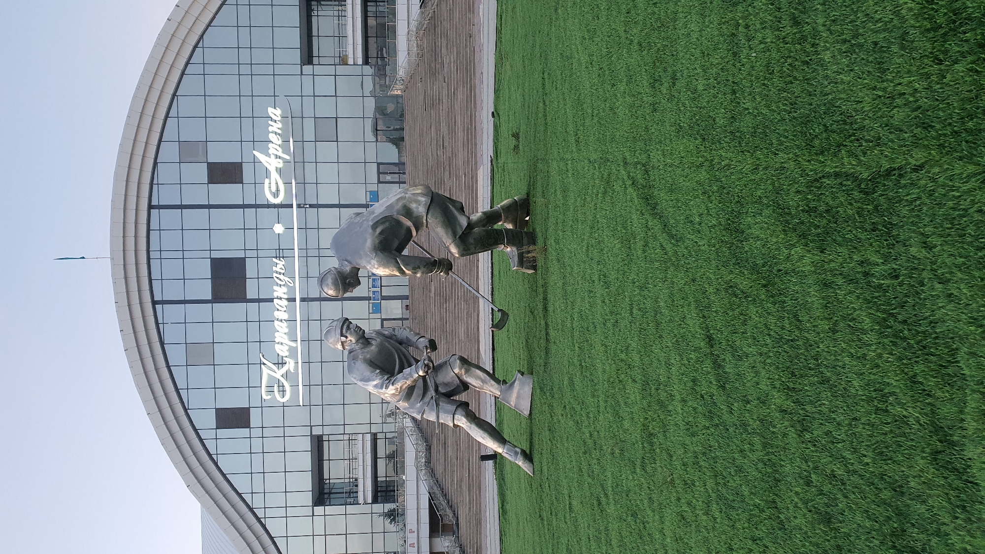 The image shows a statue of two ice hockey players in front of a large, modern building. The statues are life-sized and made of metal, depicting the players in action with hockey sticks. They are positioned on a well-maintained green lawn. The building behind them features a large, arched glass facade with the words "Караганды Арена" (Karaganda Arena) prominently displayed in illuminated white lettering. The building has multiple levels with visible windows and balconies. A staircase leads up to the entrance of the building. The sky is clear and blue, suggesting daytime. The overall scene appears to be an outdoor public space, likely a plaza or park area in front of a sports complex.