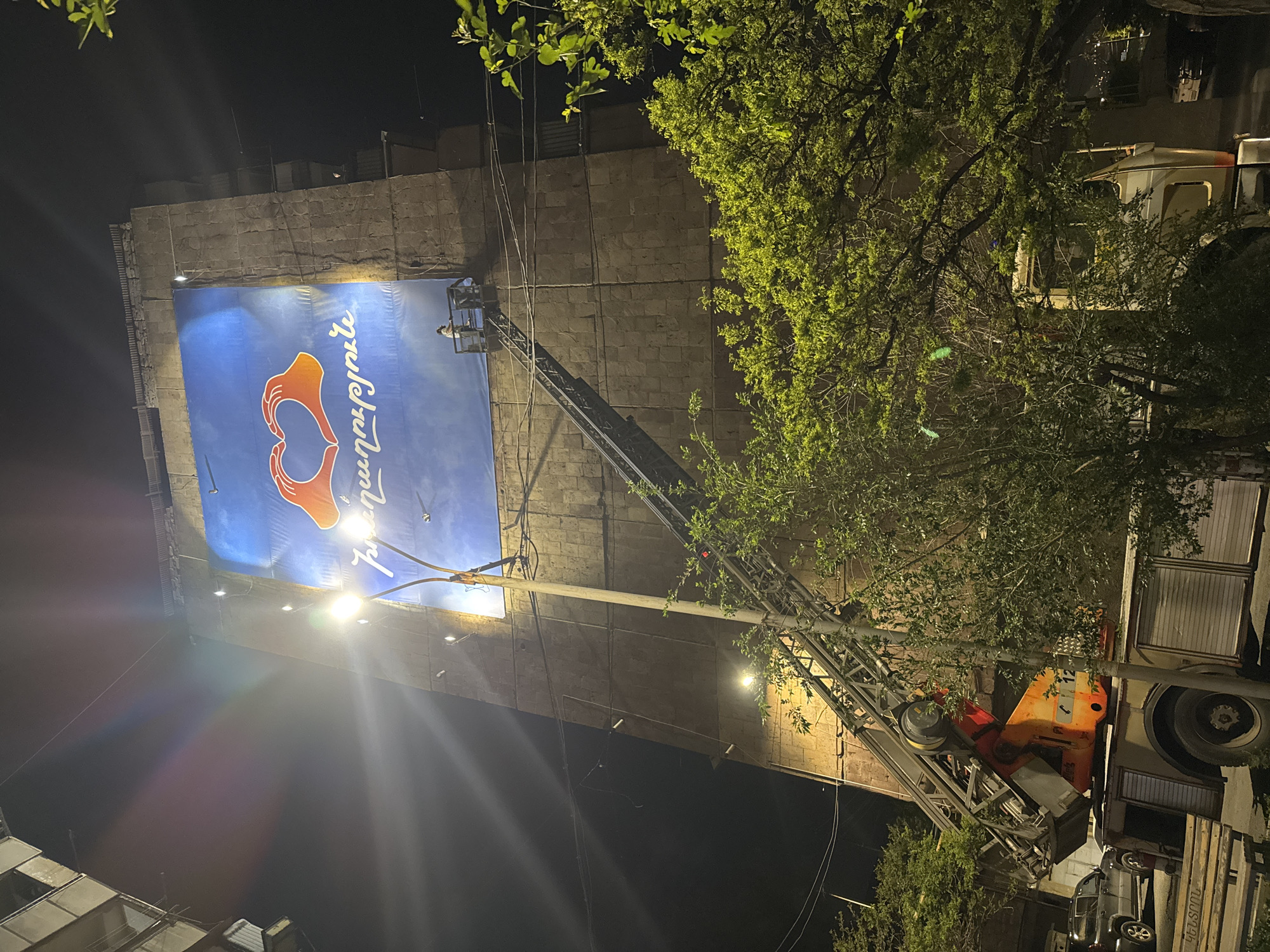 At night in Yerevan, Armenia, a large blue banner with an orange hand-heart logo and Armenian text is being installed or maintained on the side of a tall building. A construction or maintenance vehicle with a telescoping arm and a bucket at the end is positioned near the building, with a worker visible in the bucket near the banner. The scene is lit by spotlights illuminating the banner and the vehicle. Trees with green leaves partially obscure the vehicle and the building. Other buildings are visible in the background and foreground.