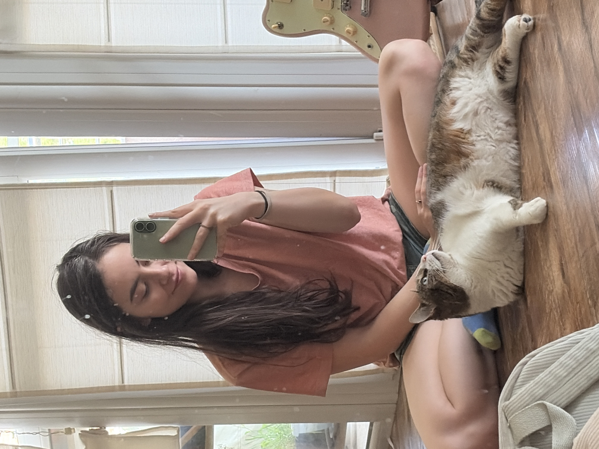 A young woman with long, dark hair is sitting cross-legged on a wooden floor, taking a selfie with her phone. She is wearing a coral-colored t-shirt and denim shorts, and a light-colored ring is visible on her right hand. A calico cat is lying on the floor next to her, looking up. In the background, blinds cover a window, and part of a pink electric guitar is visible. The scene appears to be indoors.