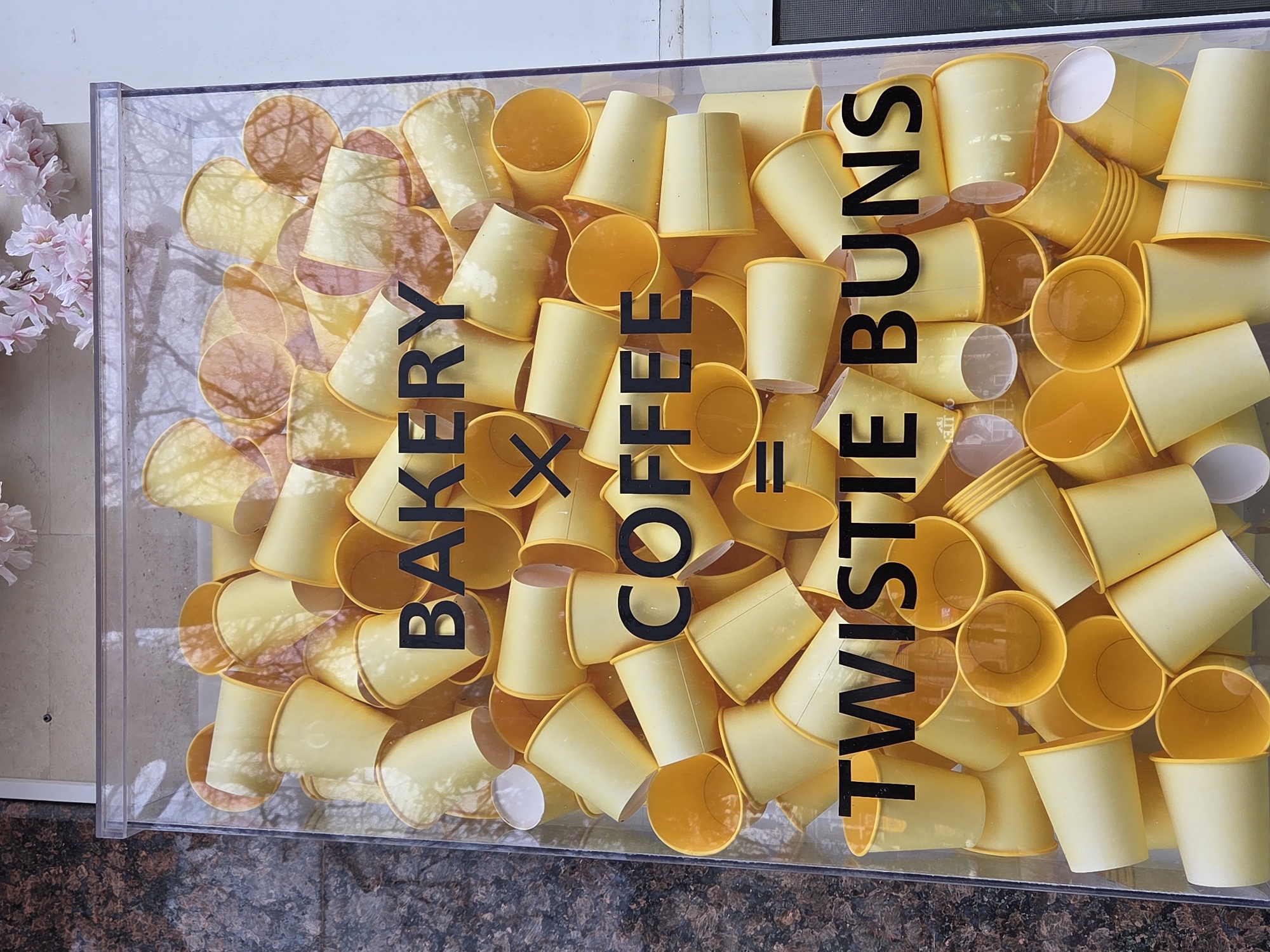 The image shows a clear, rectangular display case filled with yellow paper cups. The front of the case is printed with the words "BAKERY X COFFEE = TWISTIE BUNS" in black lettering. Some of the cups are oriented open-end up, revealing their yellow interiors, while others are upside down. The cups are densely packed, creating a textured, chaotic fill. A pink flower arrangement is visible to the left of the display case. The surface below the display case is a dark granite. Based on visual evidence, the location cannot be determined.