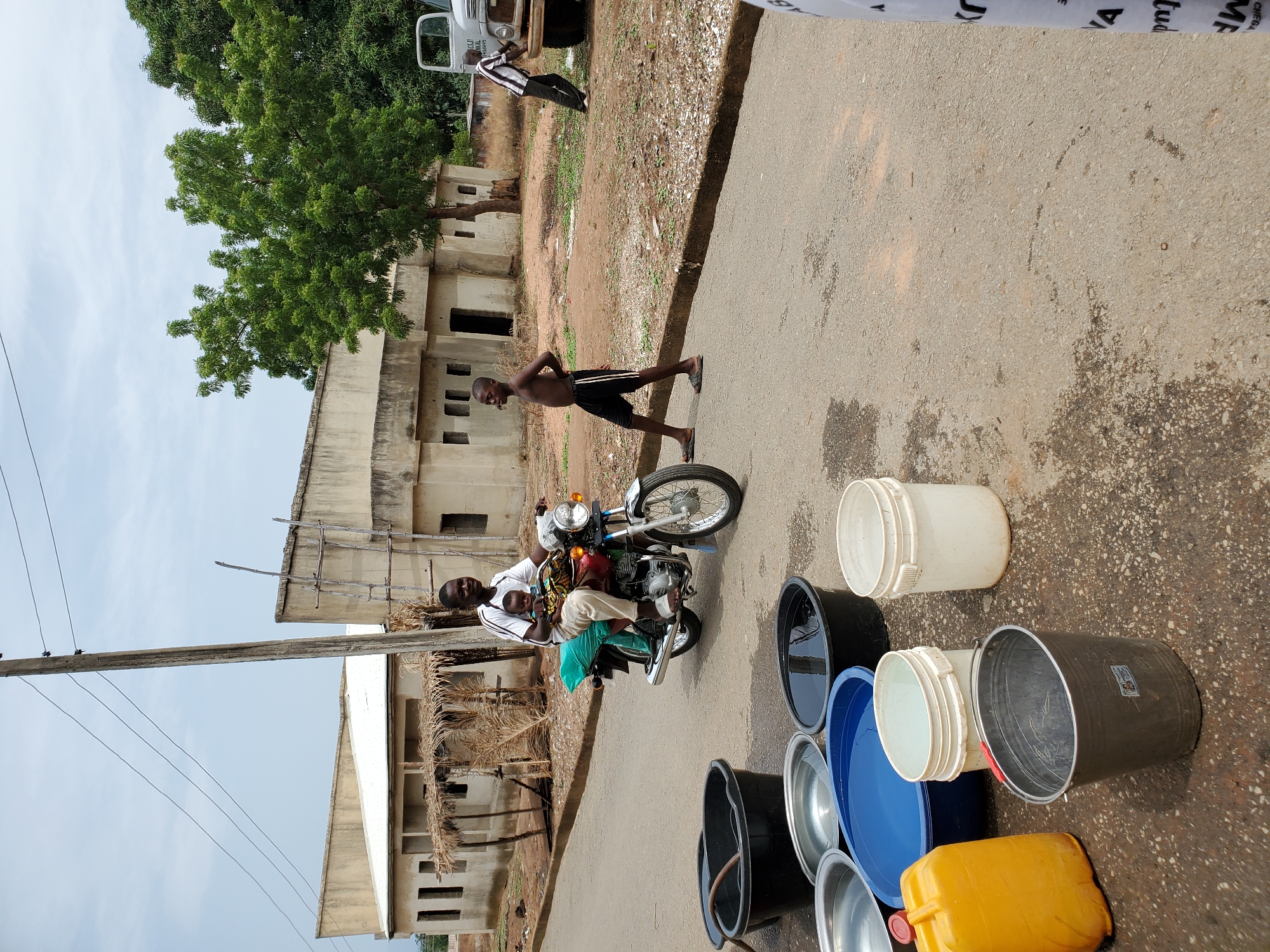 The image shows an outdoor scene in Mararaba, Nigeria. Two men are on a motorcycle on a paved road. A young boy stands to the side of the road, and an adult man is walking near a white truck. A collection of buckets and a yellow container sit on the ground near the road. Buildings with deteriorated facades are in the background, with trees nearby. Power lines are visible in the sky. The road shows signs of water or dampness.