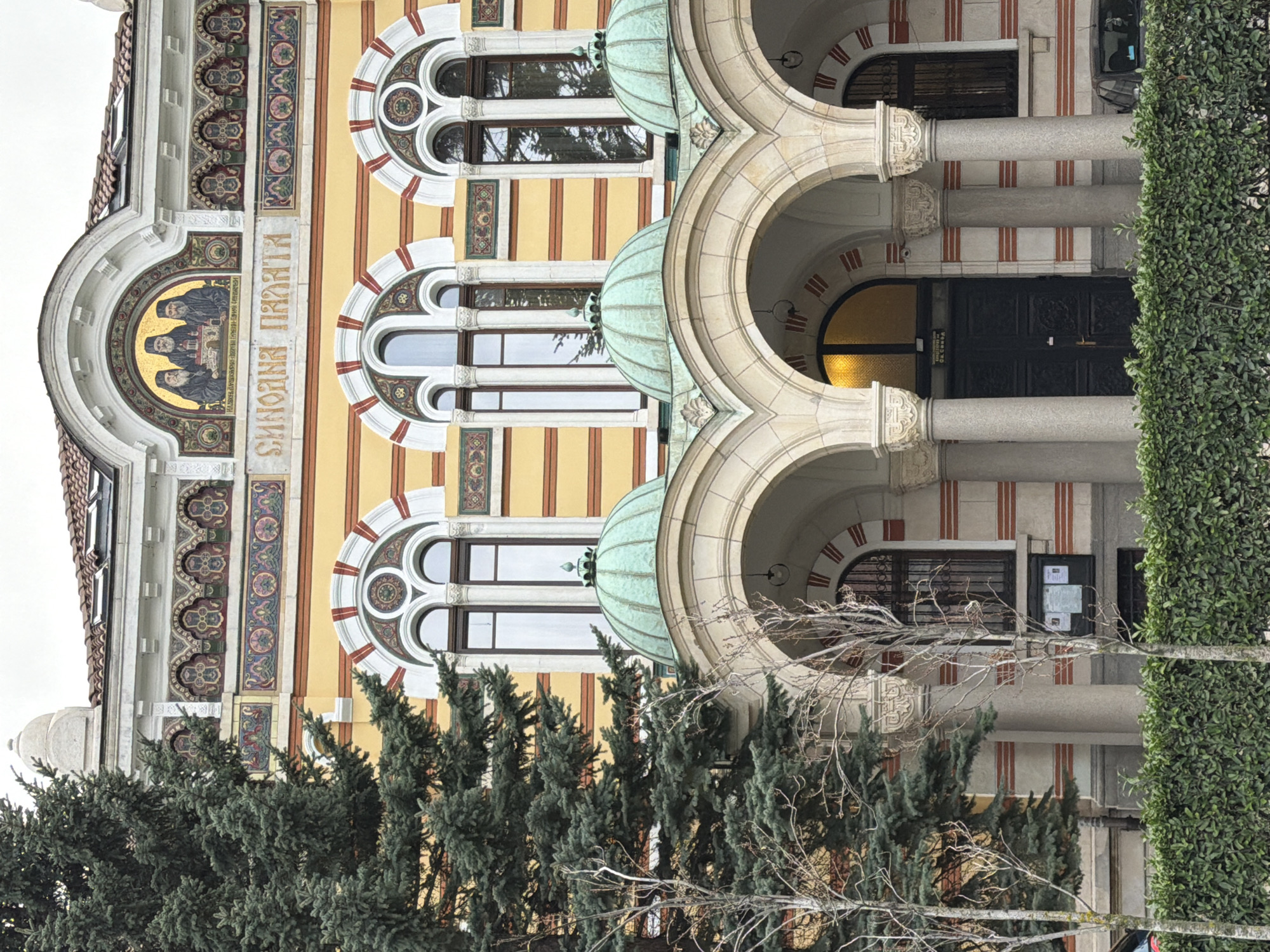 The image captures the exterior of a building featuring a mix of architectural styles, located in an unknown city. The building's facade exhibits a blend of Byzantine and Art Nouveau elements.
A prominent mosaic adorns the upper section of the building, depicting three figures in dark robes. An inscription in Cyrillic script flanks the mosaic. The building is constructed with yellow walls accented by vertical red stripes and features arched windows with circular designs. The building incorporates a series of arches supported by columns, creating a portico.
A dark wooden door with metal ornamentation is positioned at the building's entrance. A well-maintained hedge borders the right side of the frame, and coniferous trees obscure the lower portion of the building's facade. A barren tree with bare branches is visible against the building's architecture. The sky appears overcast, suggesting cloudy weather.