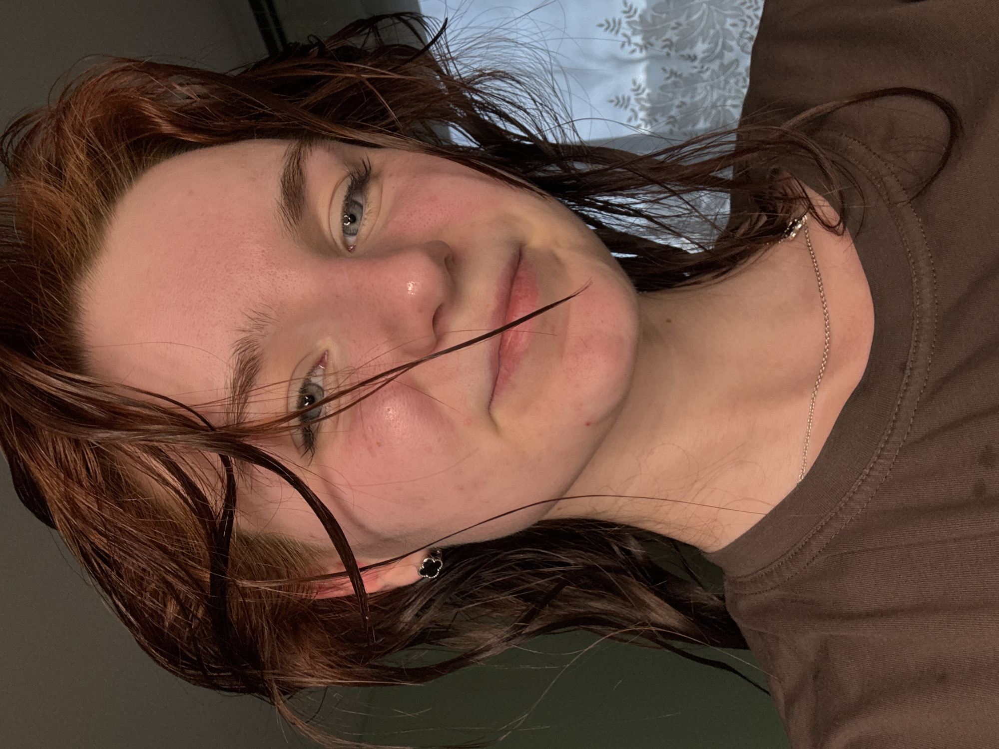 The image is a close-up selfie of a young person with wet, reddish-brown hair. The person has fair skin and grey eyes. A single strand of hair is across the person's face. The person is wearing a brown shirt and a silver necklace. A black earring is visible in the person's left ear. The background is indistinct, with a white, patterned curtain visible. The location and city in Russia are not determinable from the image.