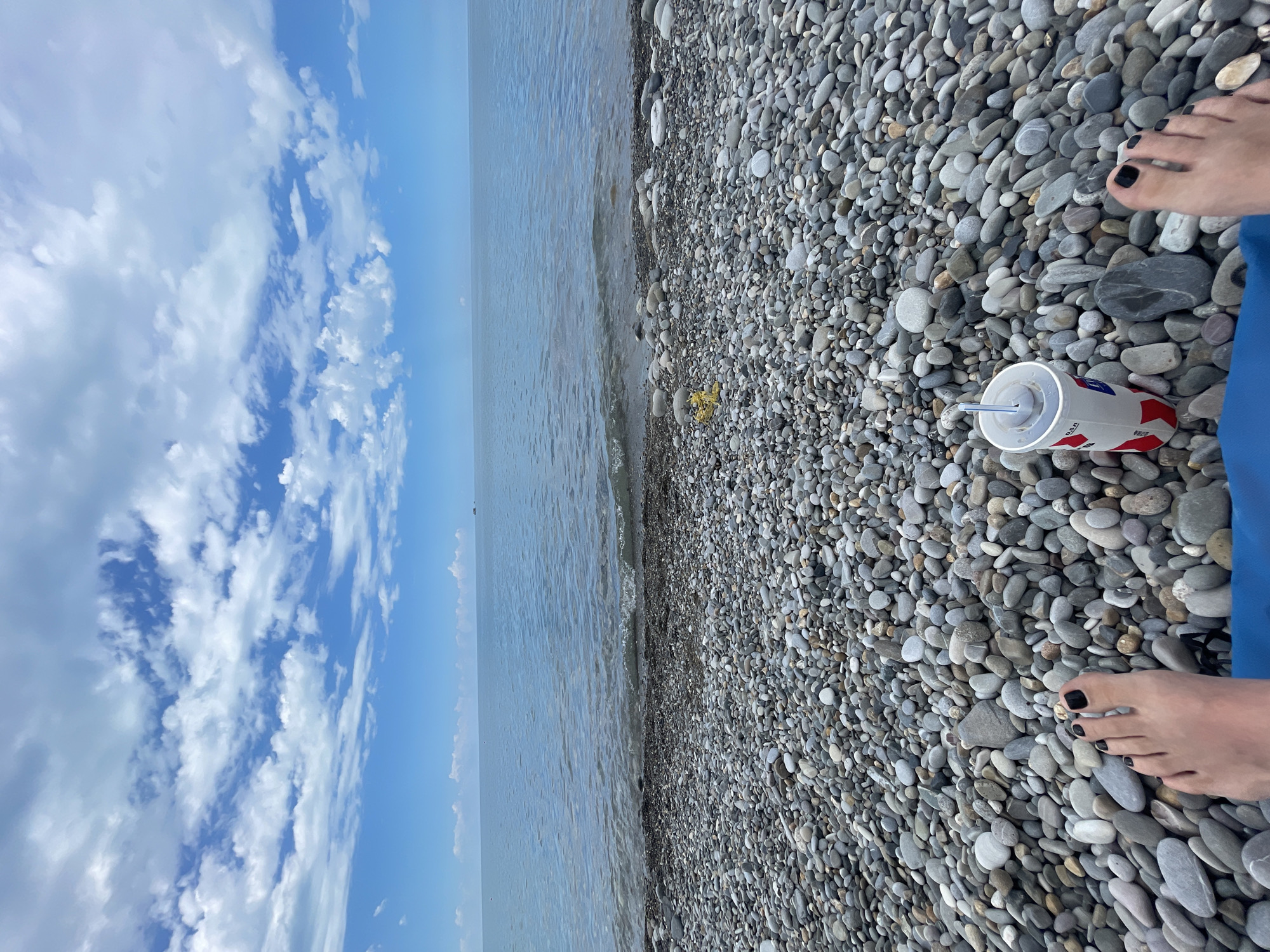 The image shows a first-person perspective of someone sitting on a pebble beach, facing the sea. In the foreground, the person's feet are visible, with black nail polish on the toes. To the right of the feet is a white cup with red and blue accents, containing a beverage and a straw. The beach is covered in smooth, gray and white pebbles. The sea is calm and stretches out to the horizon under a cloudy blue sky. The horizon line is visible with a faint, dark object, possibly a ship or buoy, in the distance. The overall scene is one of relaxation and leisure by the sea.