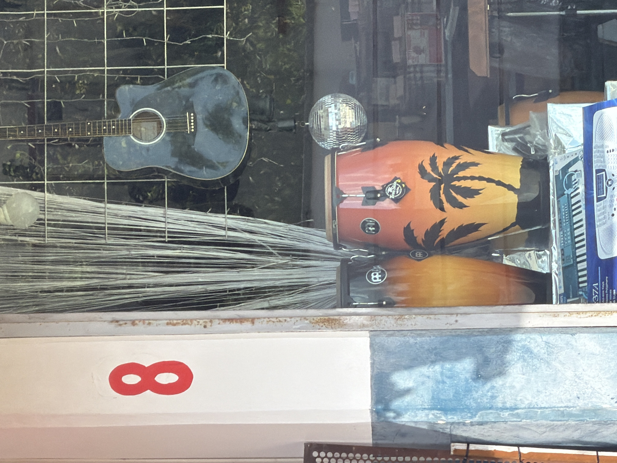 This image displays a storefront window showcasing musical instruments and related items, likely in Vigevano, Italy. The objects are arranged horizontally within the window, though the photograph itself is rotated vertically.
On the left, a dark blue acoustic guitar is prominently displayed. To its right, a small, silver disco ball hangs above a set of two bongos or congas. These drums feature an eye-catching orange-to-yellow gradient design, adorned with black silhouettes of palm trees, suggesting a tropical theme. A circular logo, possibly a stylized "B" or "BB," is visible on the upper drum. Further to the right, a portion of a white box containing what appears to be a keyboard or synthesizer is visible, with the text "37A" clearly printed on its side.
Below the guitar and extending towards the center of the display is a large bundle of fine, white, string-like material, possibly part of a decorative element or a large brush. The window reflects elements from the outside environment, including what looks like a dark background with some grid patterns, possibly from a security gate or window frames. Below the window display, on a white wall, a large red number "8" is painted. A rusted metal strip runs horizontally along the top of this wall section, just beneath the window. The overall scene is a static commercial display, likely from a music store.