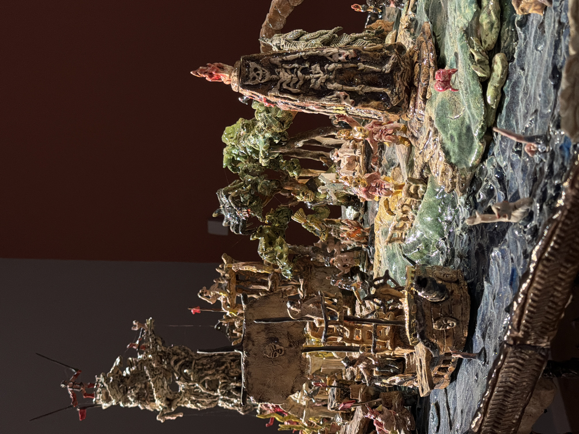 The image depicts a miniature diorama, likely an art installation, featuring a chaotic scene with a pirate ship and a monstrous, skeletal tower. The diorama is situated in Tel-Aviv, Israel, though the specific location within the city is not provided.
In the foreground, a weathered pirate ship with a tattered sail bearing a skull emblem is partially submerged in choppy, dark blue water. Several small, humanoid figures, possibly pirates or mythical creatures, are scattered across the ship's deck and in the water. Some figures appear to be in motion, with arms raised or in poses suggesting struggle or action.
Dominating the mid-ground and background is a grotesque, towering structure that resembles a skeletal figure encased in dark, textured material. It is adorned with a skull motif and a fiery red element at its peak. Lush, stylized green trees with bulbous foliage surround this structure, creating a surreal, jungle-like environment.
Scattered around the base of the tower and on small, rocky islands are numerous small, crudely sculpted figures. These figures exhibit a range of colors, primarily earthy tones, pinks, and reds, and are depicted in various poses, some appearing to be in conflict or distress. A few figures are submerged in the water, with only limbs visible.
On the left side of the diorama, a tall, twisted structure reminiscent of a rocky outcrop or a makeshift tower rises, topped with a figure holding two swords. Red flags are visible on several of the structures and on the ship, adding pops of color to the otherwise dark and ominous scene. The overall impression is one of a macabre and fantastical battle or event. The lighting suggests an indoor setting, possibly a museum or gallery, with a dark background and focused illumination on the diorama.