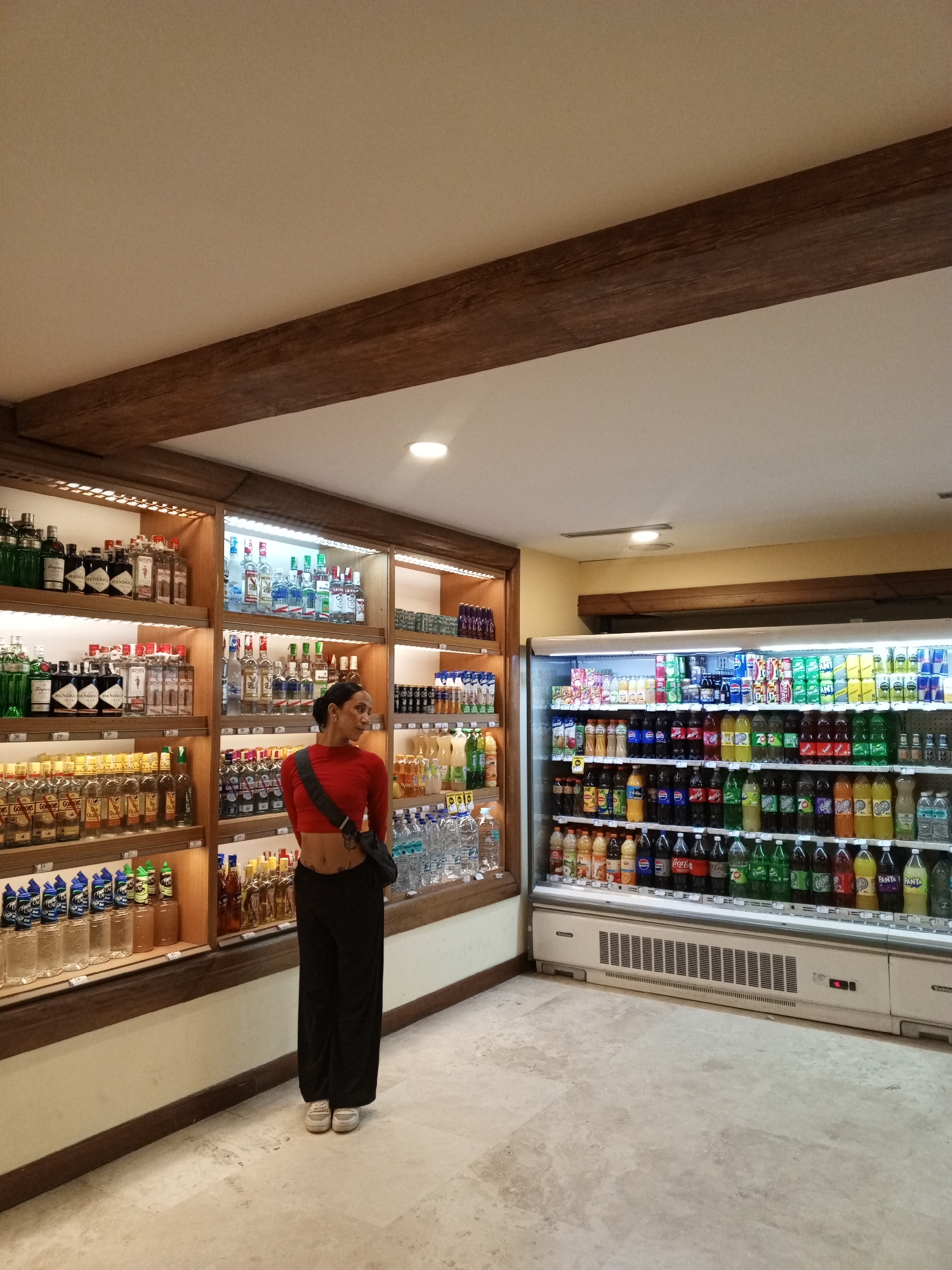 A full-length shot captures a woman standing in an aisle of a store. She has dark hair pulled back, wears a red long-sleeve crop top, black pants, and white sneakers. A black strap bag is slung across her body. She is looking to her left, towards shelves stocked with liquor bottles. To the right of the liquor shelves is a refrigerated display filled with various beverages. The location is possibly in the city of Porlamar, Venezuela. The store features wooden beams along the top of the liquor shelves and the ceiling, with neutral-colored walls and a tiled floor.