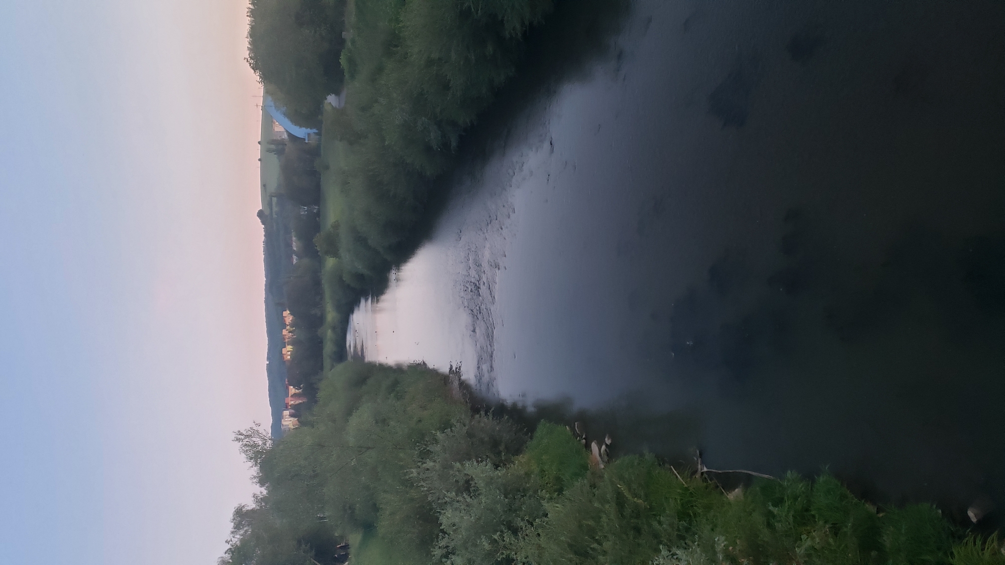 This is a high-angle, vertical shot of the Torysa River flowing through Prešov, Slovakia, at dusk. The river dominates the foreground, reflecting the pale, gradient sky. On either side of the river, lush green vegetation lines the banks, with trees and bushes forming dense borders. In the distance, across the river and on rolling hills, a cluster of residential buildings, some brightly colored, are visible. To the right of the frame, a portion of a bright blue structure, possibly part of a building or infrastructure, juts out against the green landscape. The overall scene is serene and natural, with a hint of urban presence in the background.