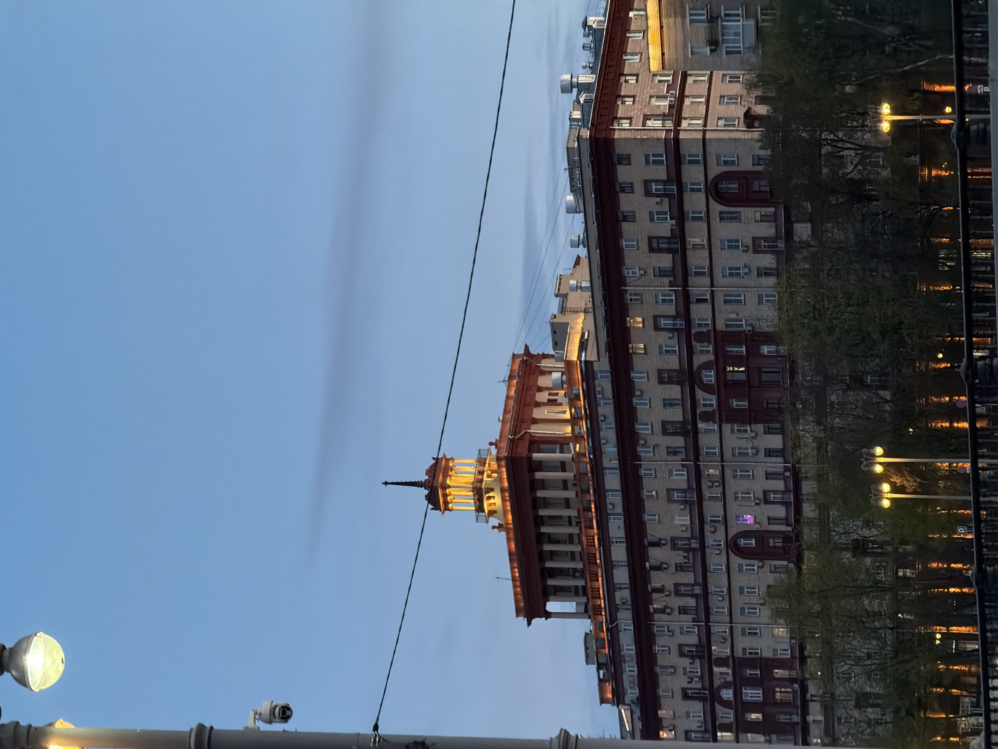 The image shows a building in Moscow, Russia, against a light blue sky. The building is tall, with a central tower-like structure featuring a spire. The building's exterior has a color scheme of light gray and red, with many windows. Below the buildings, there is a partial view of trees and street lights. There is also a utility pole in the lower left corner.