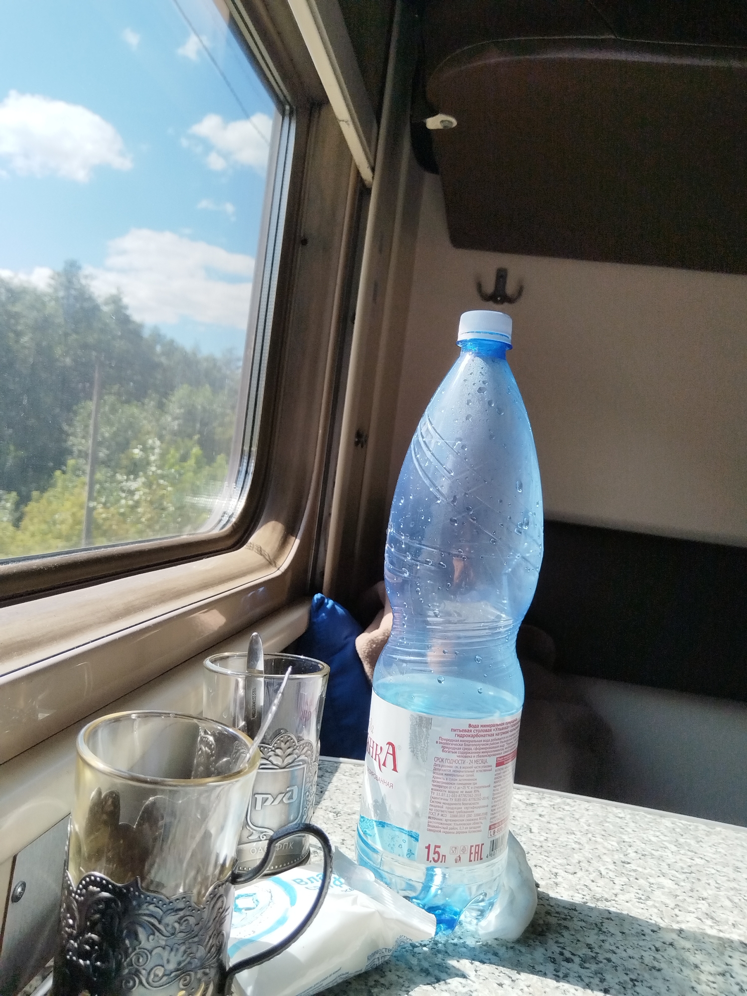 A plastic bottle of water and two glasses are on a table inside a train car, with a window showing a landscape of trees and sky outside. The bottle is partially filled with water and condensation covers its exterior. One glass is in a decorative metal holder. A blue pillow is visible behind the objects. The scene is lit by natural light coming through the window. The location is the city of Yumatovo, Russia.