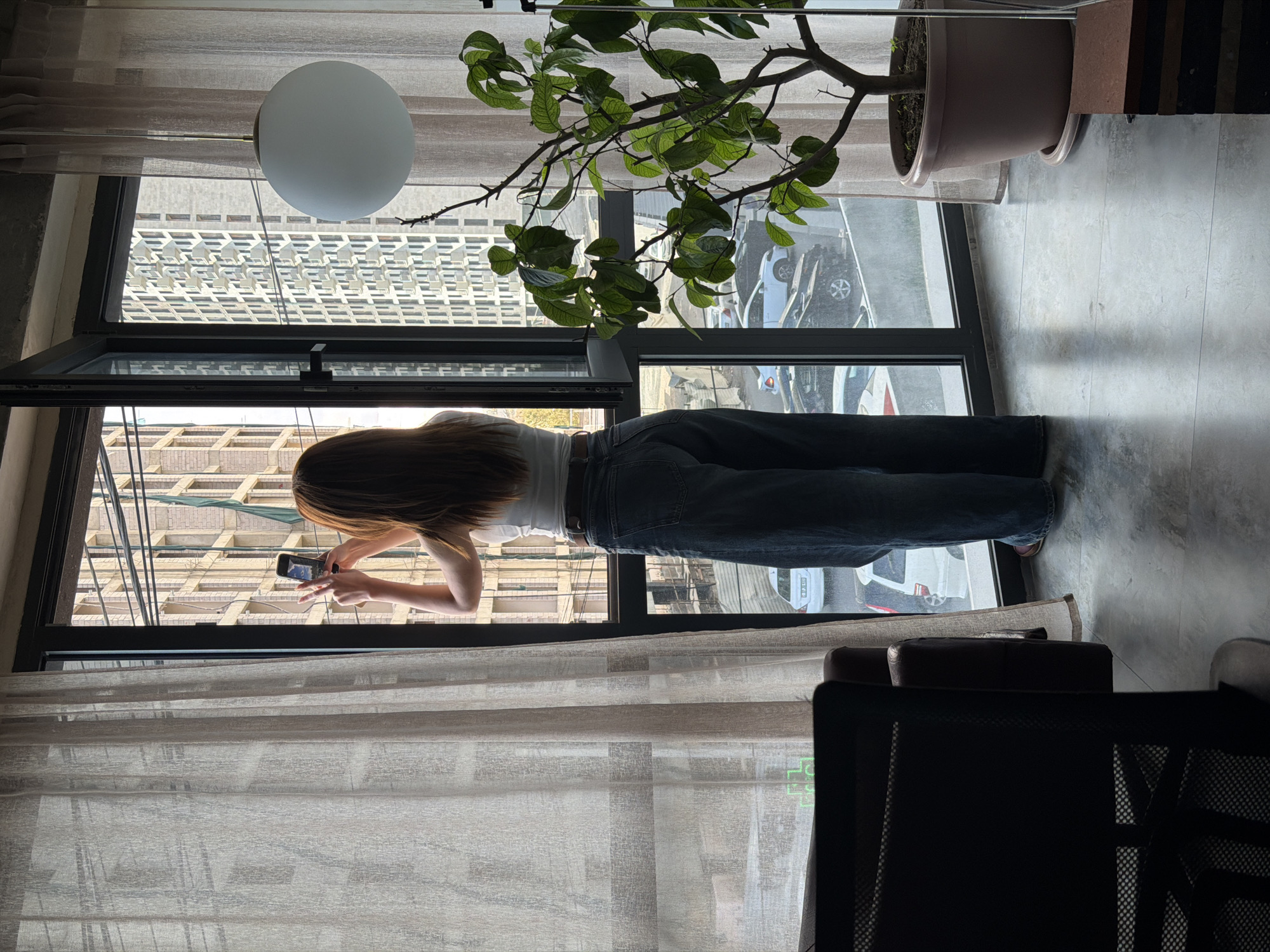 A person, viewed from behind, is photographing the cityscape of Yerevan, Armenia, from an open window. The individual is wearing a white top and dark jeans. The photograph is taken from inside a room, revealing curtains, a spherical light fixture, a potted plant, and a portion of a chair. The buildings visible through the window suggest an urban environment. The person is holding a smartphone to capture the scene outside.