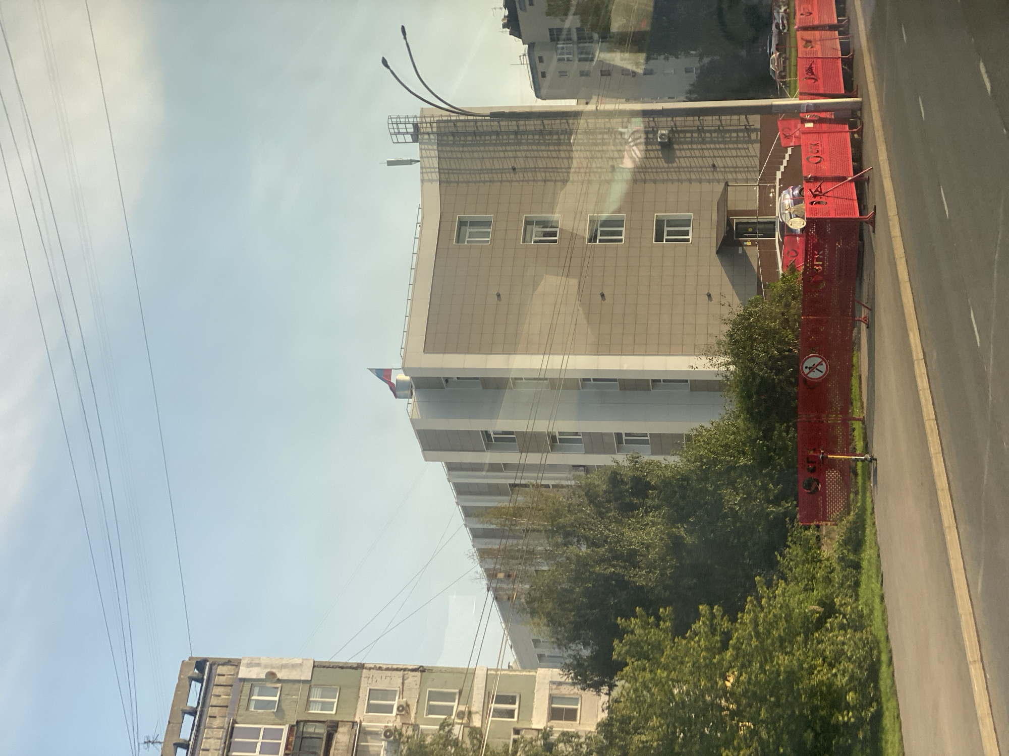 The image captures a view of a street in Kemerovo, Russia, on a clear day. On the left, a multi-story apartment building with a concrete facade and visible air conditioning units stands under a bright blue sky with scattered clouds. To the right of this building, a modern, light-colored administrative-style building with a grid-like facade and numerous windows is prominently featured. A Russian flag is visible on the roof of this building. In front of the administrative building, a red construction fence encloses an area, with a parked electric scooter and a small car visible behind it. Trees and greenery line the area between the road and the buildings. A lamppost with two curved lights is positioned near the administrative building, and power lines stretch across the sky. The road in the foreground is paved and appears to be a street in a city.