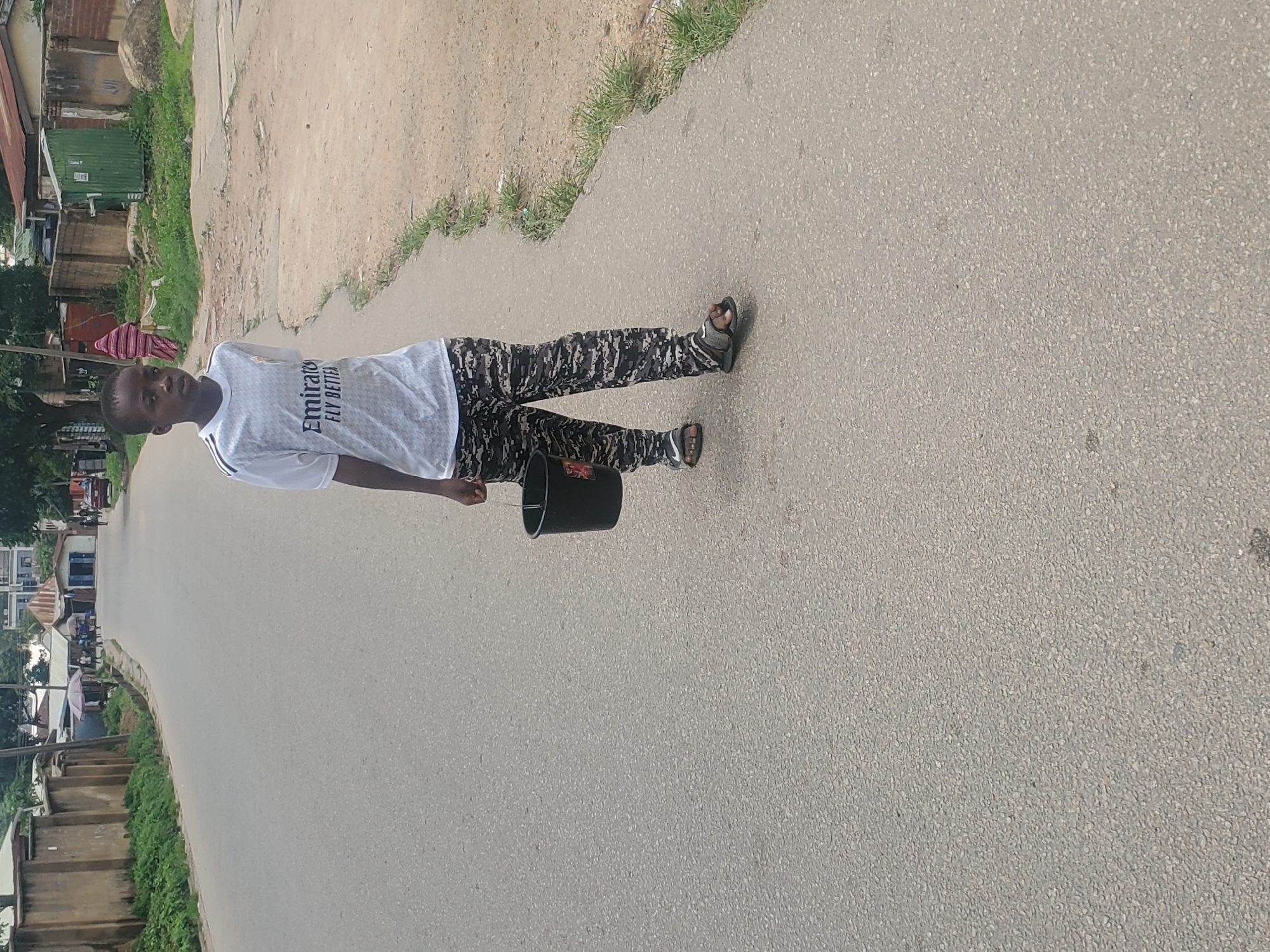 A young boy in a white t-shirt and patterned pants walks on a paved road in Jos, Nigeria, carrying a black bucket. The road appears to be in a residential area, with buildings and fences lining the sides. There are scattered trees and vegetation along the roadside. Other people and vehicles are visible in the background, suggesting a typical urban scene. The sky is overcast.