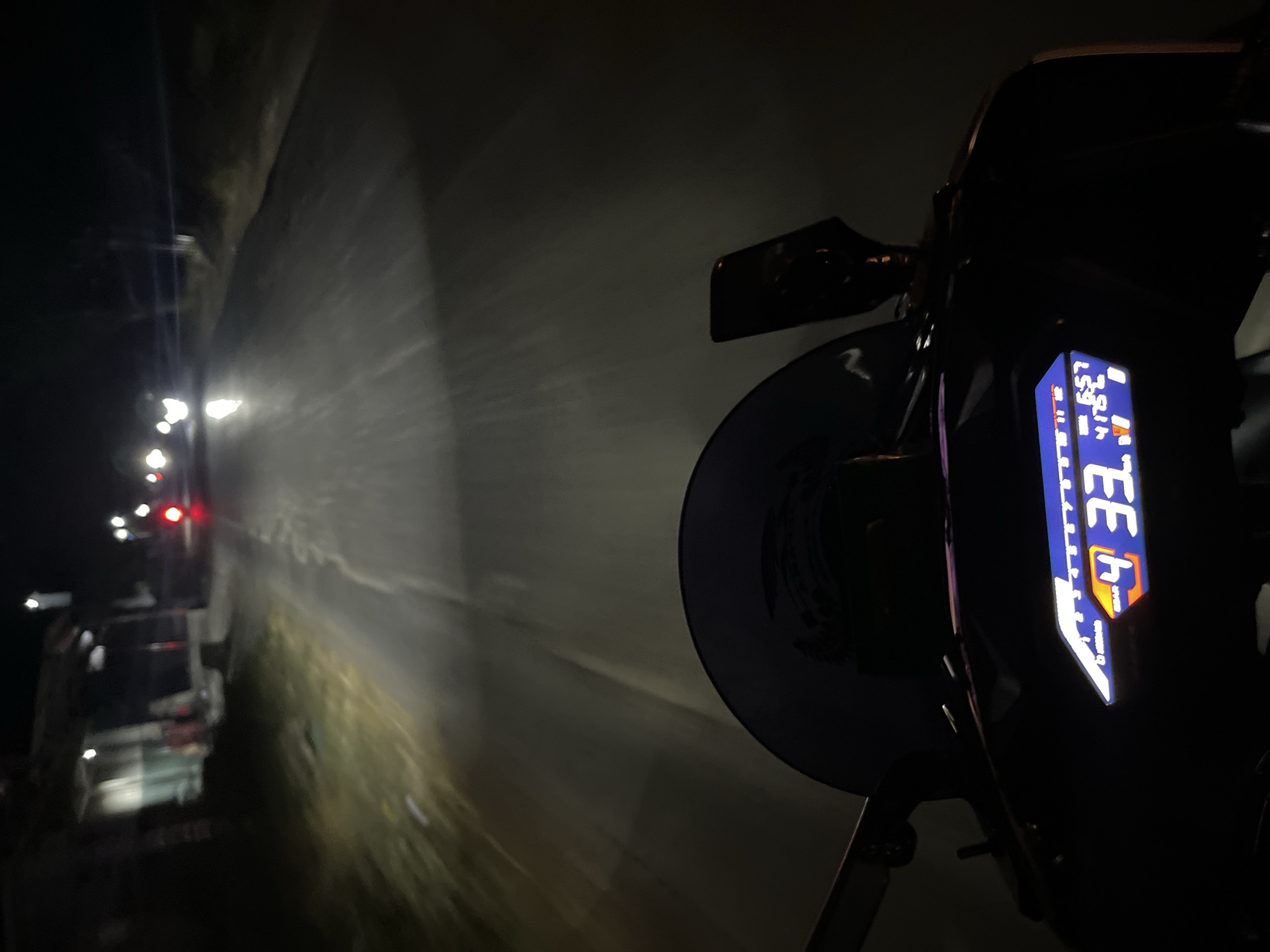 The image is taken from the perspective of a motorcycle rider at night on a paved road in Pecangaan, Indonesia. The motorcycle's digital dashboard is visible in the foreground, displaying "GEAR 4", a speed of "33", and other indicators. The road ahead is illuminated by the motorcycle's headlights, revealing a slight sheen from moisture. On the left side of the road, there are structures with lights, suggesting businesses or residences. Further down the road, the taillights of another vehicle are visible, along with oncoming headlights. The environment appears to be rural or suburban, with dark vegetation lining the sides of the road. The lighting conditions are low, characteristic of a nighttime scene.