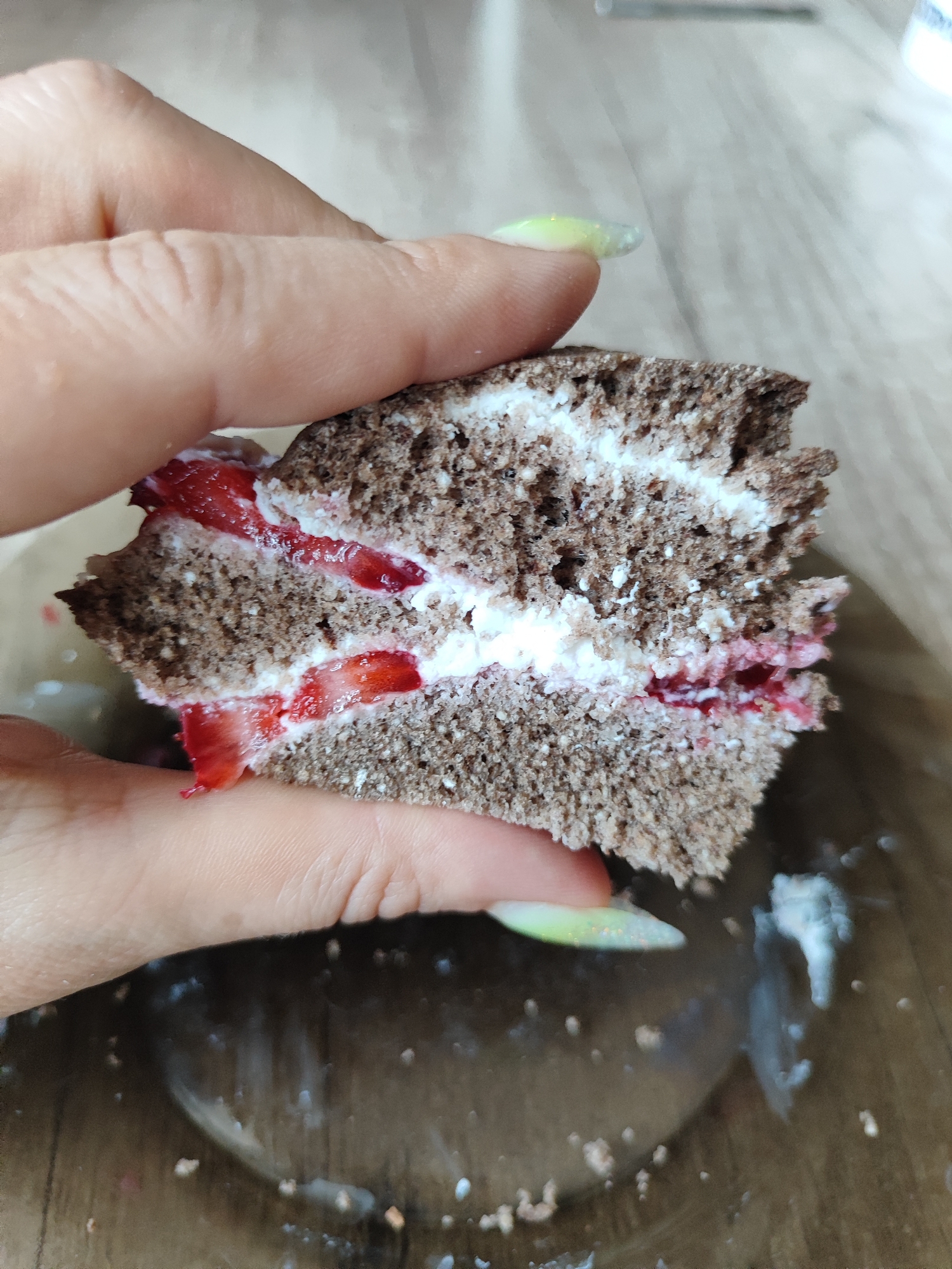 A person's hand is holding a slice of chocolate cake with white cream and strawberry filling. The cake is dark brown, with visible pores, and the cream appears to be whipped. Sliced strawberries are interspersed within the cream layers. The background is a wooden surface, with some white smudges or spills visible. The scene is captured in a close-up shot, focusing on the cake and the hand. No specific location in Almaty, Kazakhstan is discernible from the image.