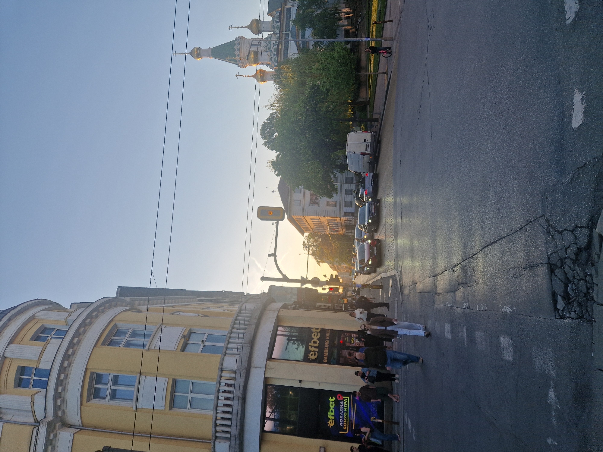 Here is a description of the image:
The image depicts a street scene in the city of Unknown, Unknown, at daytime. On the left is a multi-story building with arched details and a cream-colored facade. There are people walking in front of the building, near a store with the word "efbet" prominently displayed on the storefront. The street is lined with cars, and there is a traffic light. In the background, there is a building with multiple golden domes. Power lines are visible against the clear sky. The overall lighting suggests it may be late afternoon or early evening. The street surface shows signs of wear and tear.