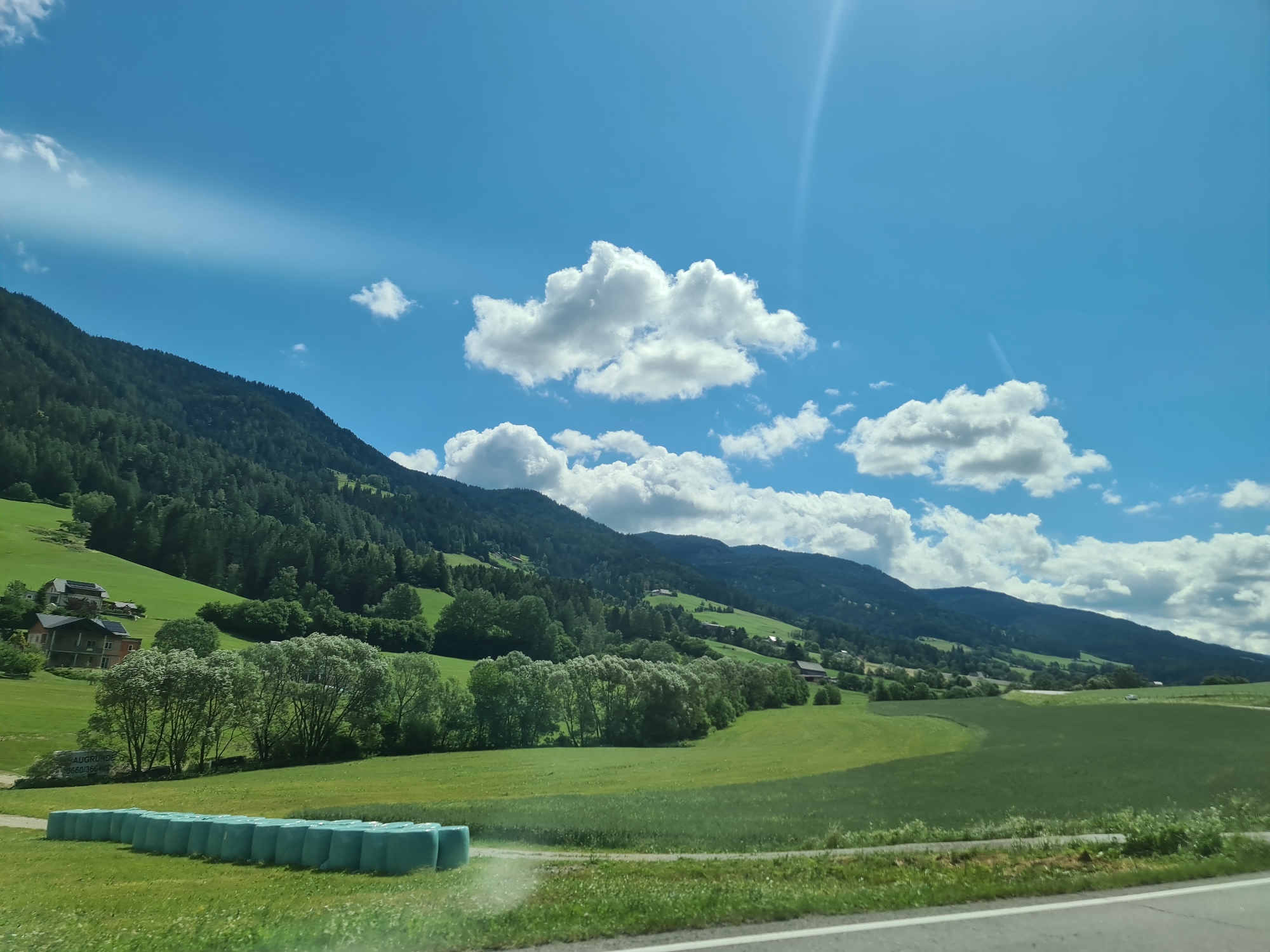 A sunny day over a lush green landscape in Scheifling, Austria, features rolling hills dotted with trees and buildings, beneath a bright blue sky filled with white, fluffy clouds. In the foreground, a row of teal-wrapped hay bales sits on a green field. A paved road with white lines runs along the bottom edge of the image. The backdrop is a majestic mountain range covered in dense green forest, with scattered houses and farm buildings nestled on its slopes. The sun's rays create a bright flare in the upper right corner of the sky.