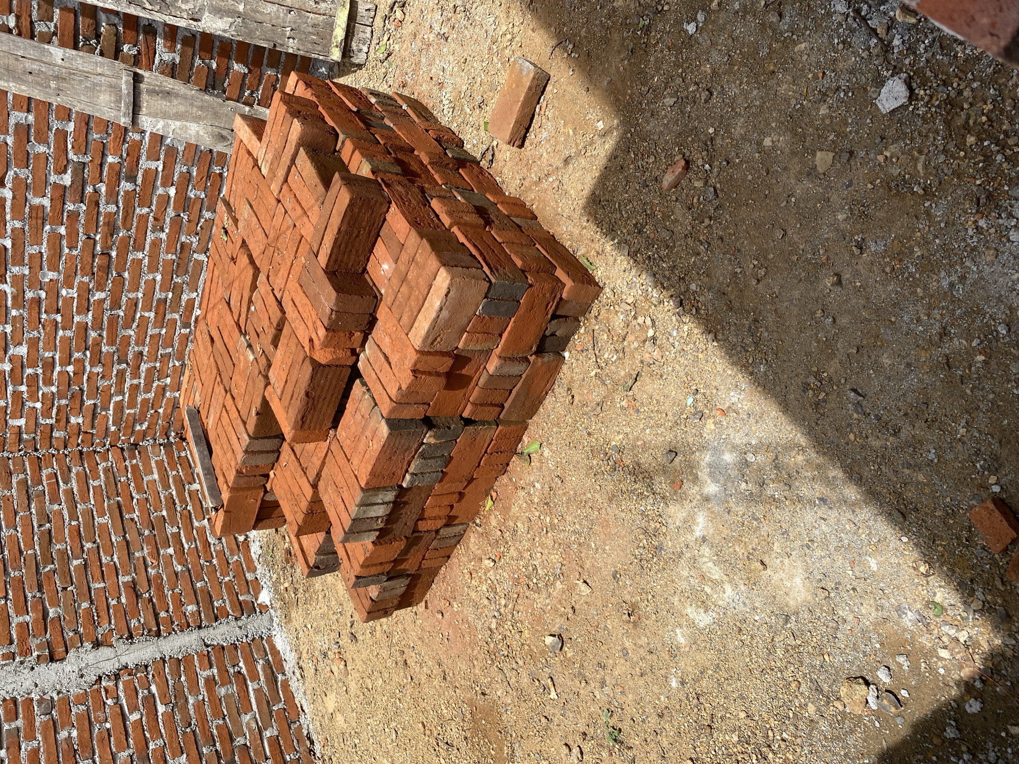 A pile of red clay bricks is stacked in a corner of a construction site. The bricks are rectangular and have a rough texture. They are stacked in layers, with some bricks placed horizontally and others vertically. The surrounding ground is covered with dirt and small stones. In the background, a brick wall is partially constructed, with visible mortar between the bricks. Two wooden planks are leaning against the wall. The scene appears to be outdoors, in a sunny environment, with shadows cast on the ground. The location is identified as Banda Aceh, Indonesia.