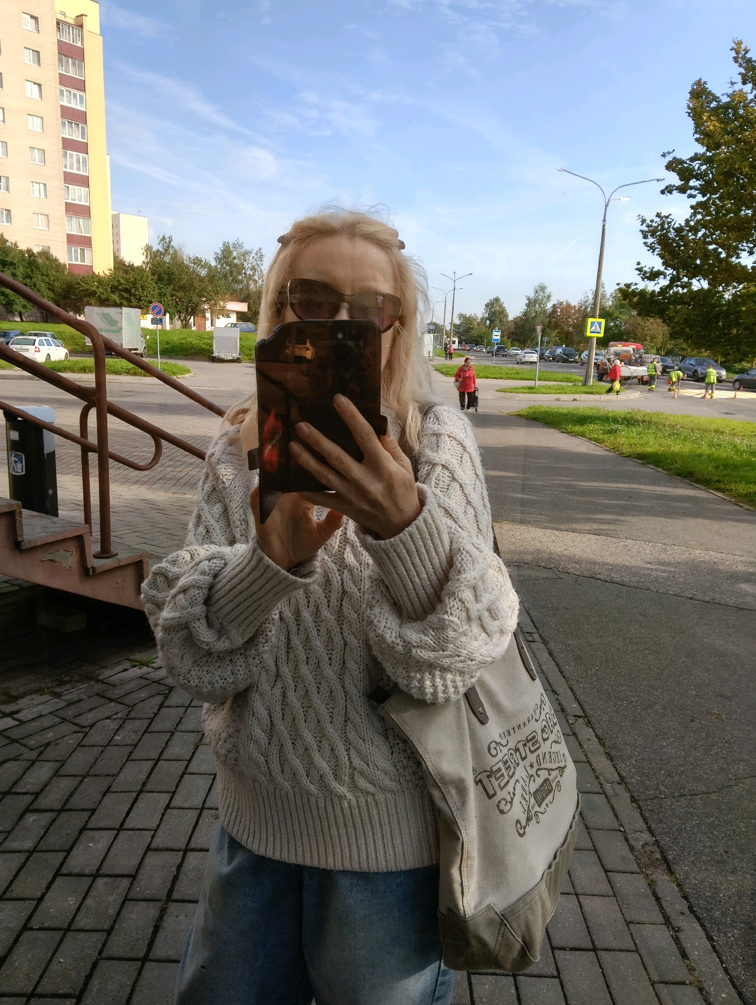 A woman with blonde hair and sunglasses is taking a selfie with a phone in her hands. She is wearing a chunky knit sweater and jeans. A large canvas tote bag is slung over her shoulder. The background shows a residential building, trees, a sidewalk, a street with traffic, and construction workers in yellow vests. The image appears to be taken in Hrodna, Belarus, based on the architecture and street signs which are not clearly visible but suggested by the context of the city.