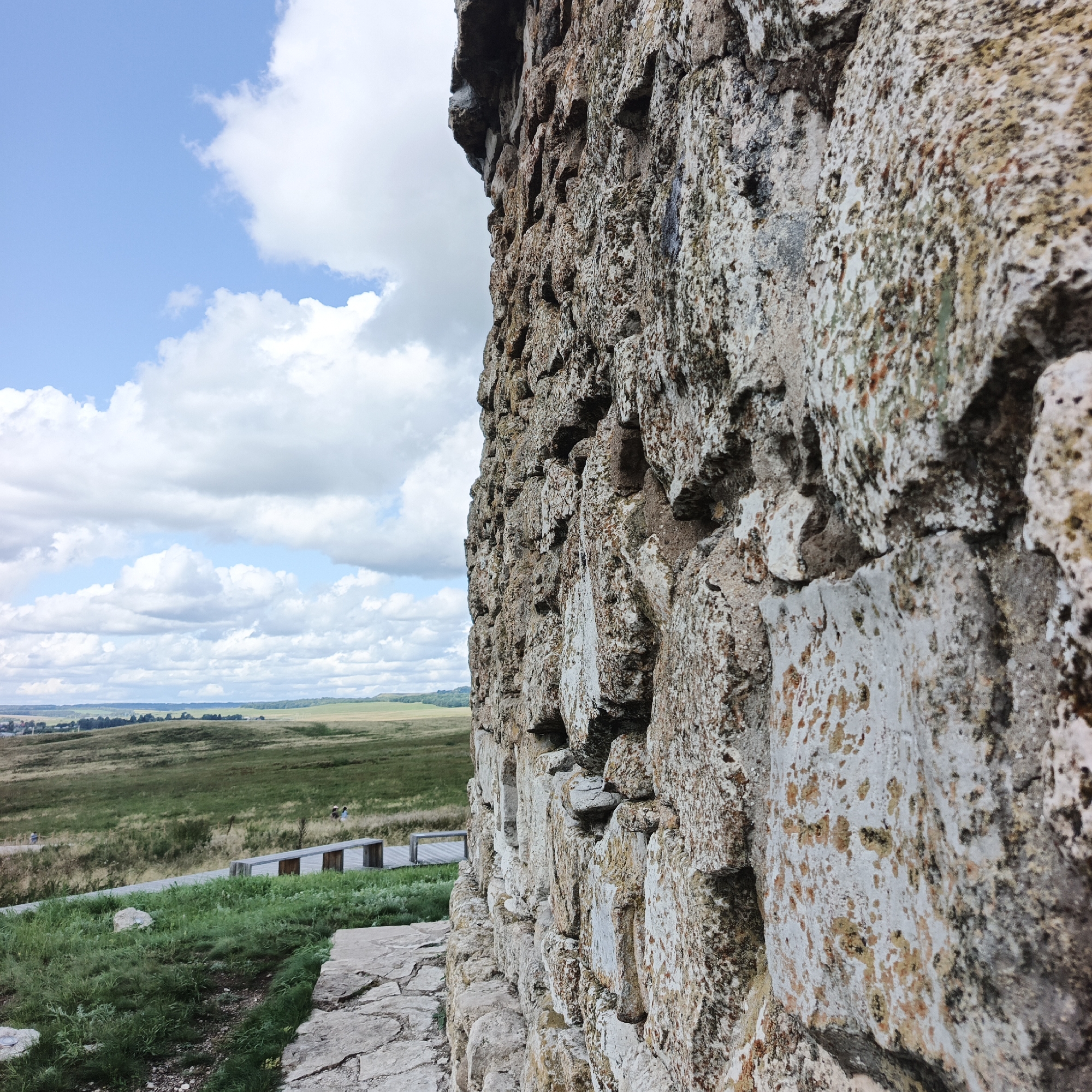 The image depicts a textured stone wall in the foreground, angled to the right, with a pastoral landscape in the background under a partly cloudy sky. The stone wall is constructed from rough-hewn, irregularly shaped stones, showing variations in color from light gray to beige and brown, with patches of green and orange lichen. The stone's surface is uneven, creating shadows and highlighting its granular texture. To the left of the wall, a path made of flat stones leads into a grassy area. Beyond this, a wooden boardwalk with benches is visible, curving through a green, rolling landscape. Further in the distance, the terrain is covered in fields of varying shades of green and brown, with a line of trees forming the horizon. The sky is a light blue, populated by fluffy white and gray clouds. A few small figures, appearing to be people, can be seen in the mid-ground on the boardwalk, suggesting a public outdoor space. The scene is likely an archaeological site or a historical monument given the presence of ruins and a preserved landscape. The location is identified as Verkhnie Termy, Russia, a place known for its historical significance.