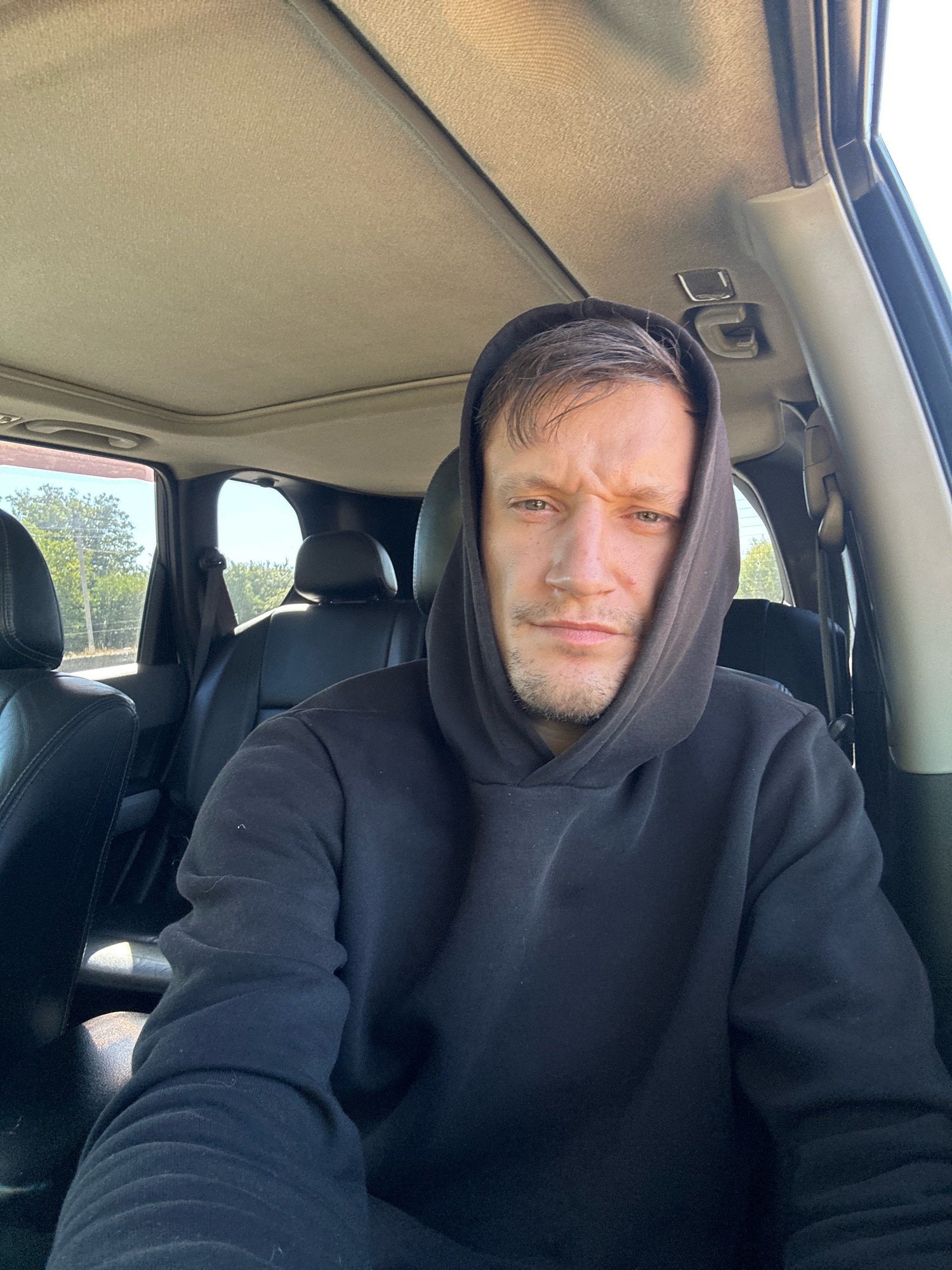 A man with light brown hair and stubble is sitting in the driver's seat of a car. He is wearing a black hoodie and has the hood pulled up. The interior of the car is visible, including black leather seats and the car's headliner. Sunlight is streaming in from the windows, illuminating the scene. Outside the car, green trees and vegetation are visible. The man is looking directly at the camera with a neutral expression. The location is the city of Makiv, Ukraine.