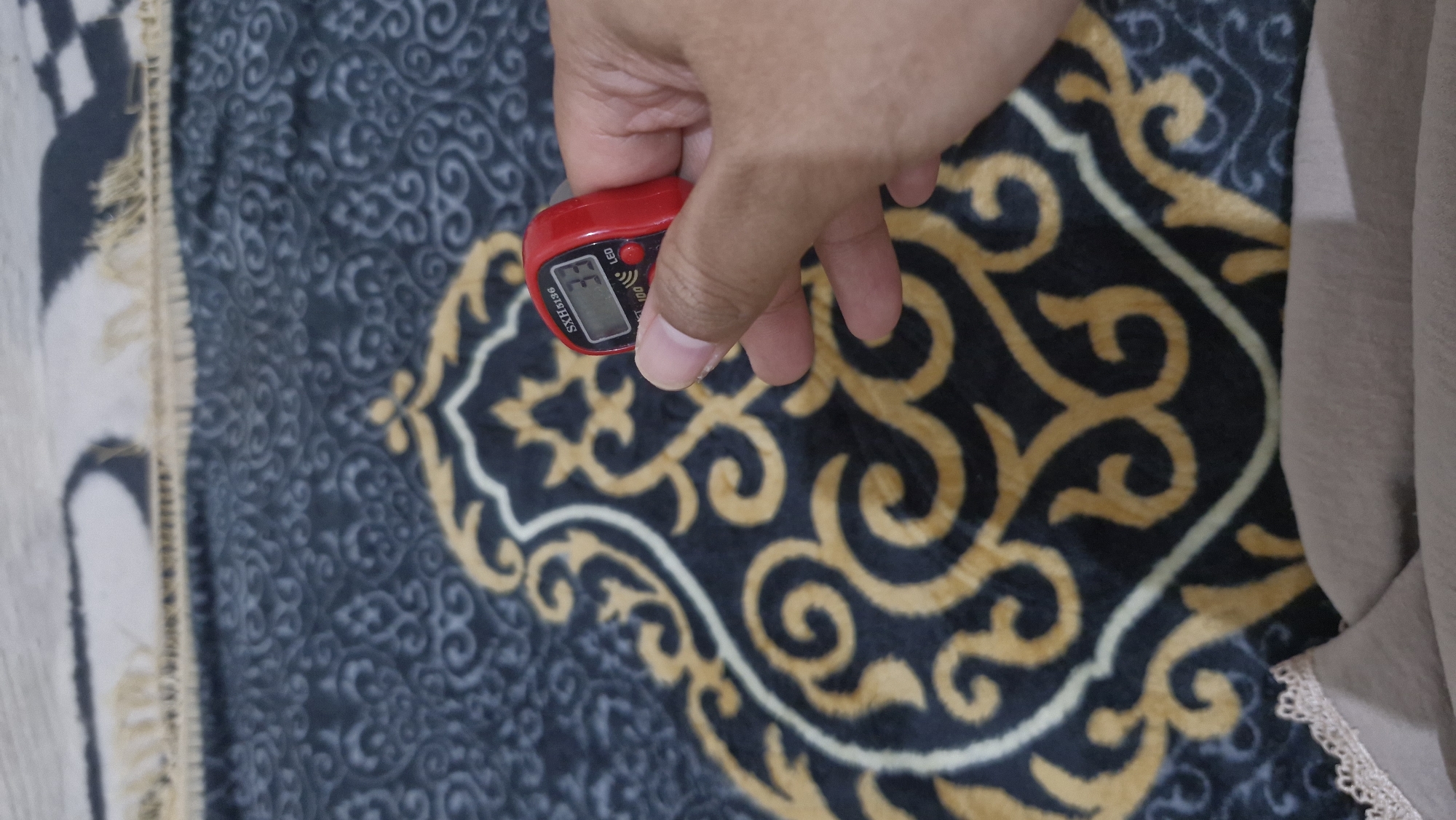A hand is holding a red digital prayer counter on a dark blue and gold patterned prayer rug. The counter displays the number 33. The rug is Islamic in design and appears to be in a home environment in Rantau, Indonesia.