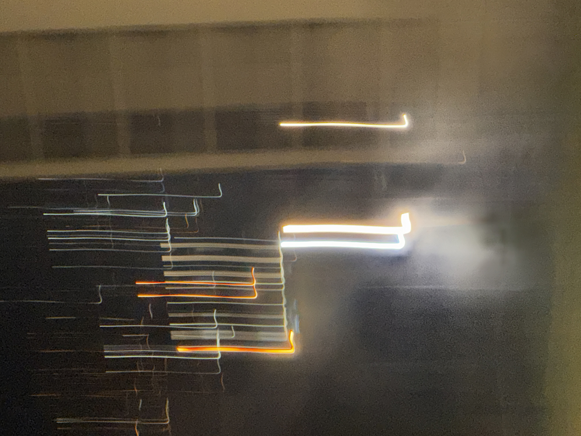 The image shows a long-exposure photograph of light trails, likely from vehicles or streetlights, against a dark, grainy background. The light trails are predominantly vertical lines of various colors, including white, orange, and yellow, with some appearing as thin, sharp lines and others as broader, blurred streaks. In the foreground, there are two prominent vertical white light trails, one slightly to the left and one to the right, with a hazy glow emanating from them. To the left and slightly higher, there are a series of thin, vertical white and orange lines that form a somewhat structured pattern, suggesting movement or stationary objects with lights. The background appears to be a building facade, rendered in muted tones of brown and gray due to the low light conditions and long exposure. The overall impression is abstract and impressionistic, capturing the motion and glow of urban light at night. No specific landmarks of Tbilisi, Georgia are discernible in this abstract depiction of light trails.