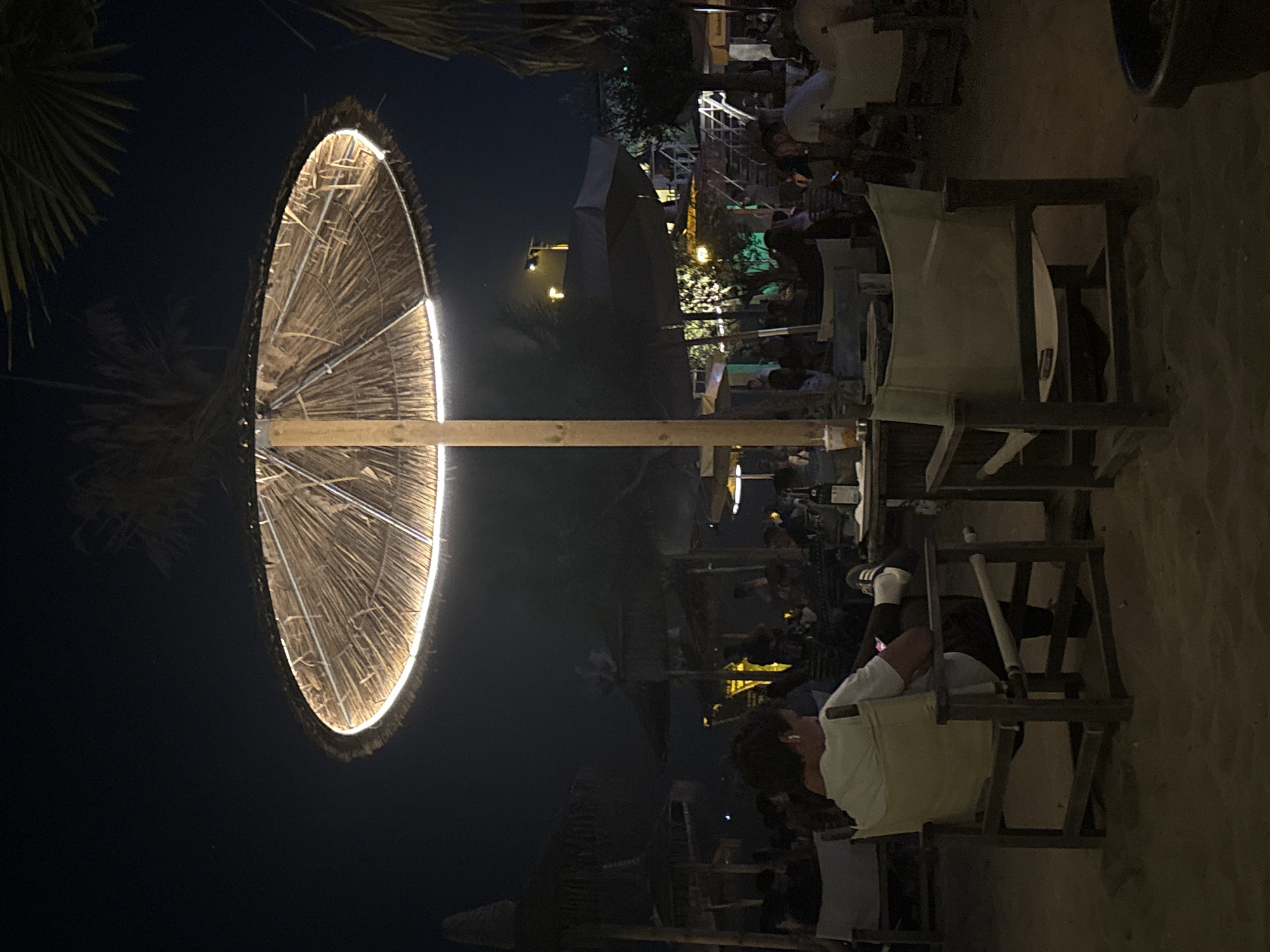 The image depicts a nighttime scene at a beachside establishment, likely a bar or restaurant, in an unknown location. The foreground features a person seated in a canvas director's chair, facing away from the camera, engrossed in their phone. They are wearing a white t-shirt and dark pants, with athletic shoes. To their right, another empty director's chair is visible. A wooden table with a bottle and glasses is situated between the chairs.
Dominating the scene is a large, straw-covered umbrella with integrated lighting, casting a warm glow. In the background, numerous other umbrellas, tables, and chairs are scattered across the sandy ground, with several patrons visible. Palm fronds can be seen against the dark night sky, suggesting a tropical or subtropical setting. The overall atmosphere is relaxed and social, with people enjoying an evening outdoors.