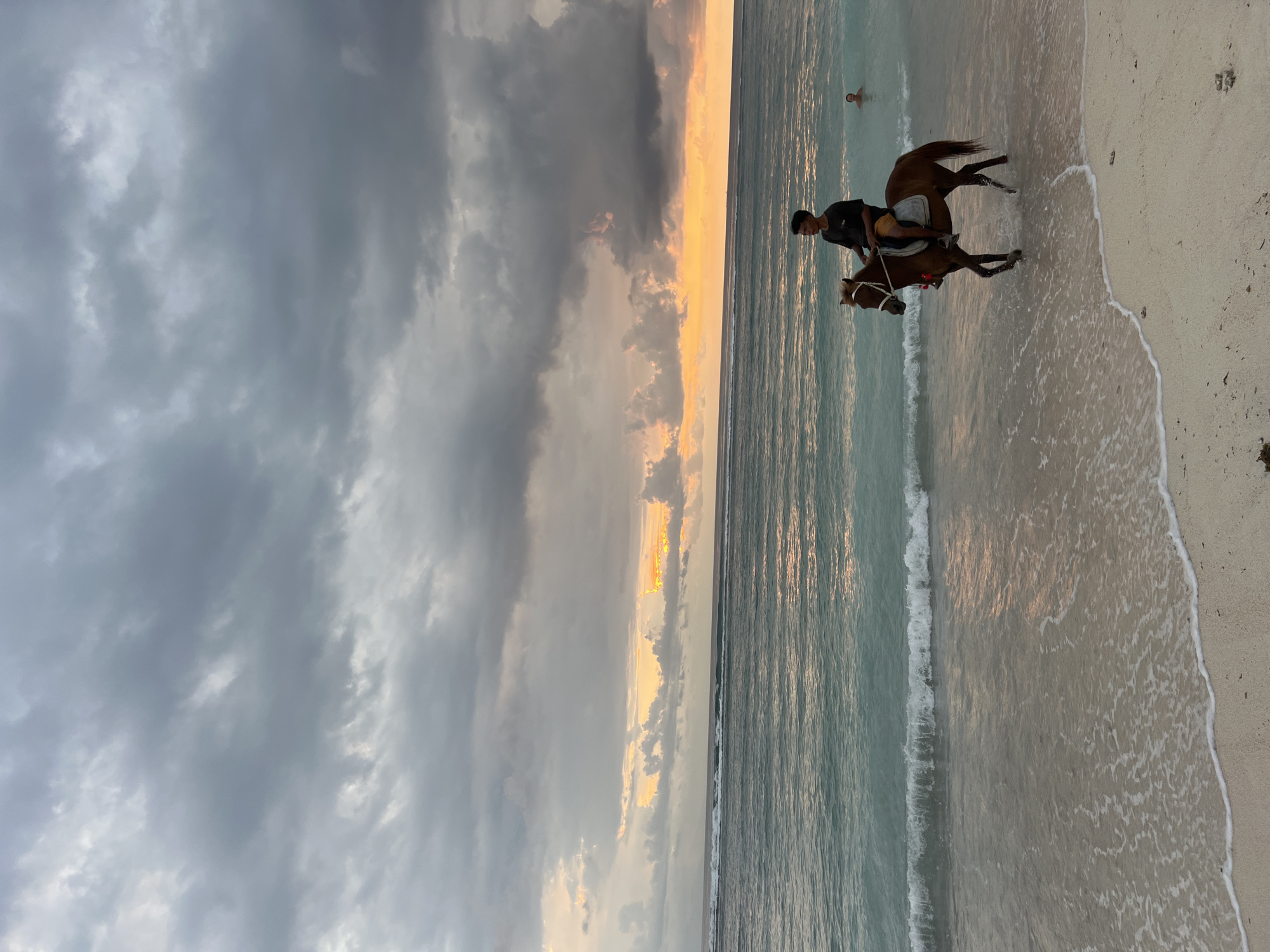A person rides a horse on a beach, presumably in the city of Unknown, Indonesia. The horse is brown, and the rider is wearing dark clothing. The ocean is visible in the background, and the sky is cloudy with orange and yellow hues indicating sunset. Another person is standing in the water in the background. The horse's hooves are splashing in the water near the shoreline, and the tide is receding. The sand is light beige in color.