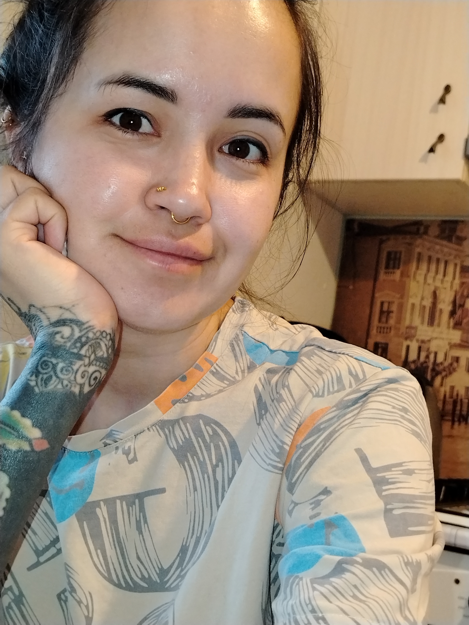 A close-up shot captures a fair-skinned woman with brown eyes and dark hair loosely pulled back. She is smiling slightly, with her left hand supporting her chin, showcasing a detailed tattoo on her wrist and forearm. She has two gold piercings in her nose. She is wearing a patterned shirt in cream, grey, orange, and light blue. A framed picture of a building sits in the background. The picture is not identified as being in the city of Noviy, Russia.