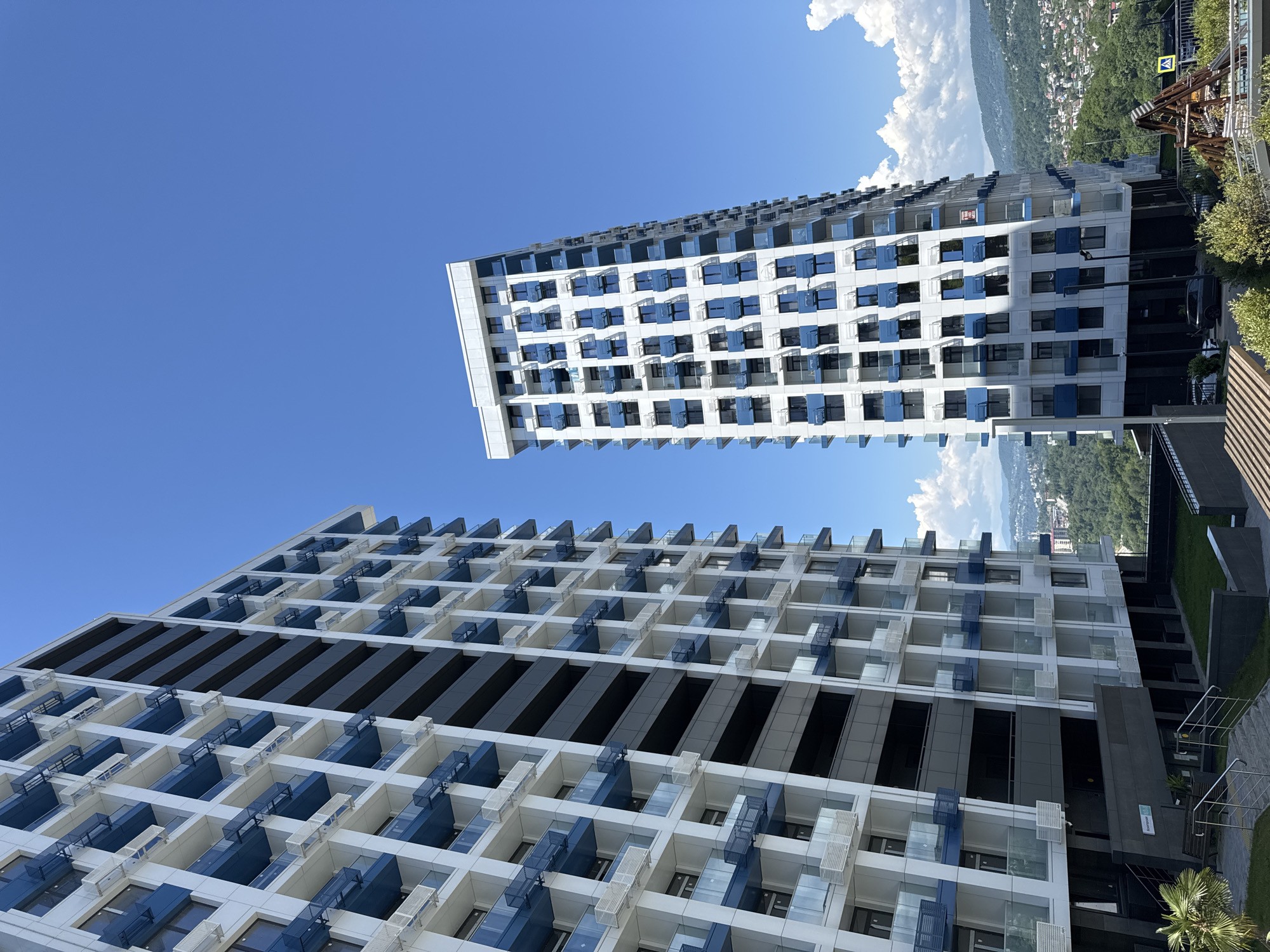 The image captures a sunny, clear day featuring two modern, high-rise buildings set against a vibrant blue sky with scattered white cumulus clouds. The architecture of both buildings is characterized by a dominant white facade, complemented by horizontal bands of blue balcony railings and transparent glass balustrades.
The building on the left, appearing taller and closer to the viewer, features a prominent central vertical section clad in dark gray panels. Its numerous balconies are arranged in a grid-like pattern, many equipped with white exterior air conditioning units. At the base of this building, a landscaped area with a small palm tree and a set of stairs leading to an entrance are visible. A dark gray panel near the ground level clearly displays Cyrillic text: "Гостиница" (Gostinitsa), which translates to "Hotel" or "Guest House."
The building on the right, positioned further in the background and appearing slightly shorter, shares the identical architectural style of white walls and blue-accented balconies. It is situated on a hillside, overlooking a sprawling landscape of rolling green hills covered in dense forestation, interspersed with residential developments and smaller buildings typical of a city. In the far right mid-ground, a wooden play structure and a yellow diamond-shaped road sign with a black arrow are partially visible.
The overall scene, with its modern resort-style architecture, prominent "Hotel" signage, and the characteristic hilly, green topography, identifies the location as the **Grand Garden (Гранд Гарден) aparthotel complex in Sochi, Russia.** The complex is known for its multi-tower design and elevated position offering panoramic views of the city and surrounding mountains.