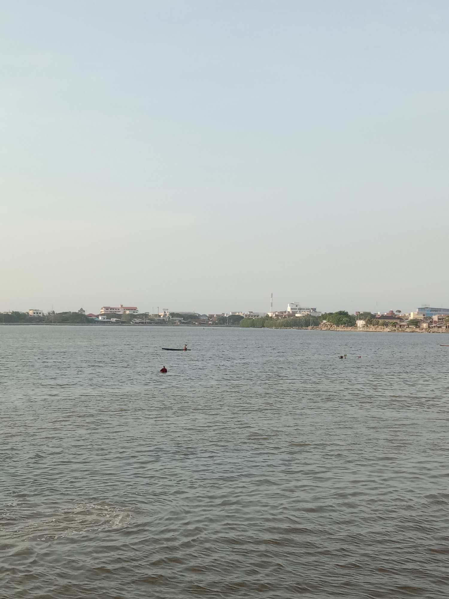 The image captures a view of the sea in Lhokseumawe, Indonesia. The city's buildings and greenery line the distant shore. In the water, a person paddles in a small boat or kayak and another person swims, marked by a floating red buoy nearby. The sky is overcast.