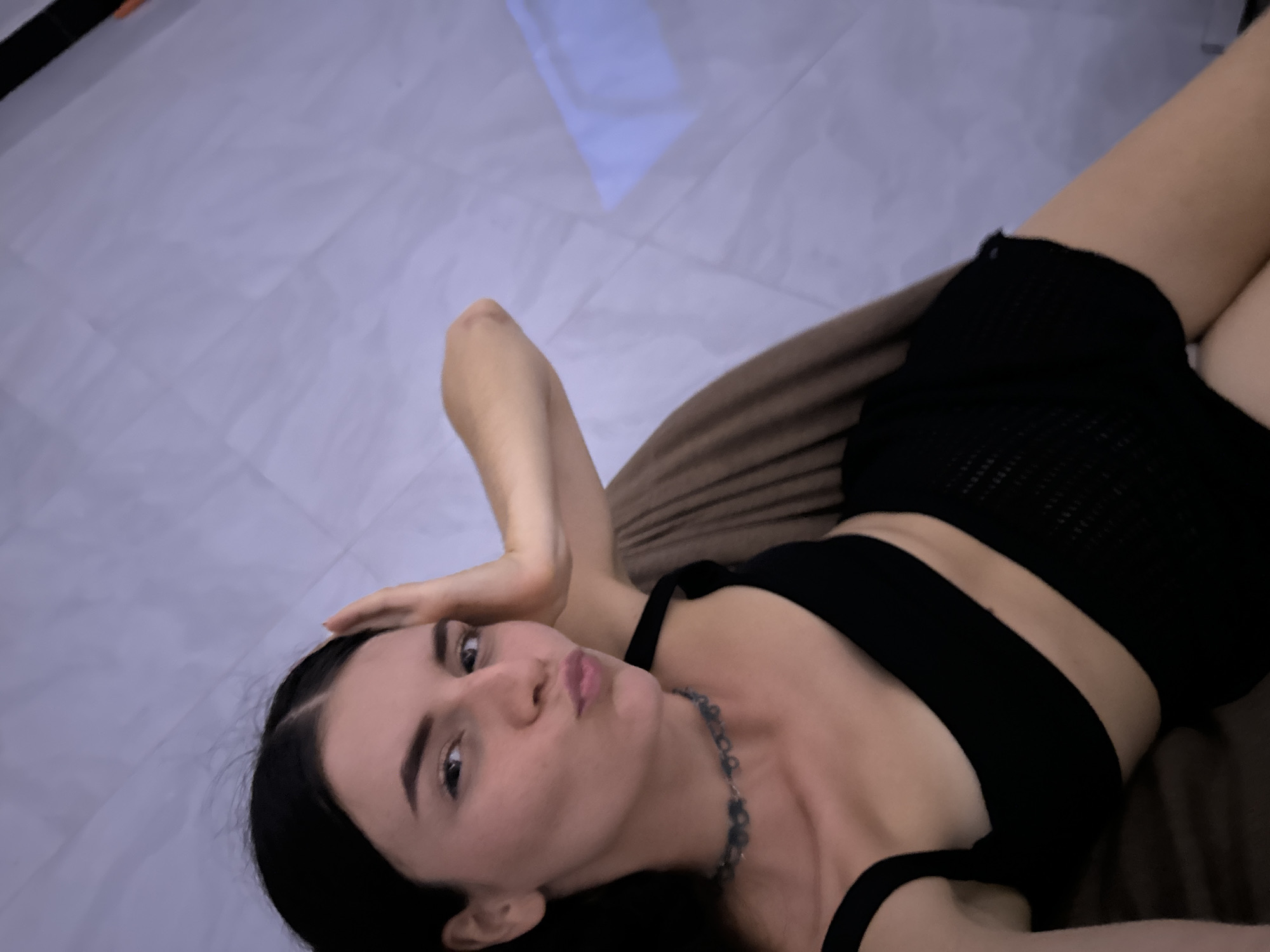 A woman lies on a brown cushion in an indoor setting. She wears a black crop top and black shorts, and a necklace. Her dark hair is loose, and she looks at the camera with her hand touching her forehead. The background includes a tiled floor and a white object on the floor. The location is not discernible as Mũi Né, Vietnam based on the image.