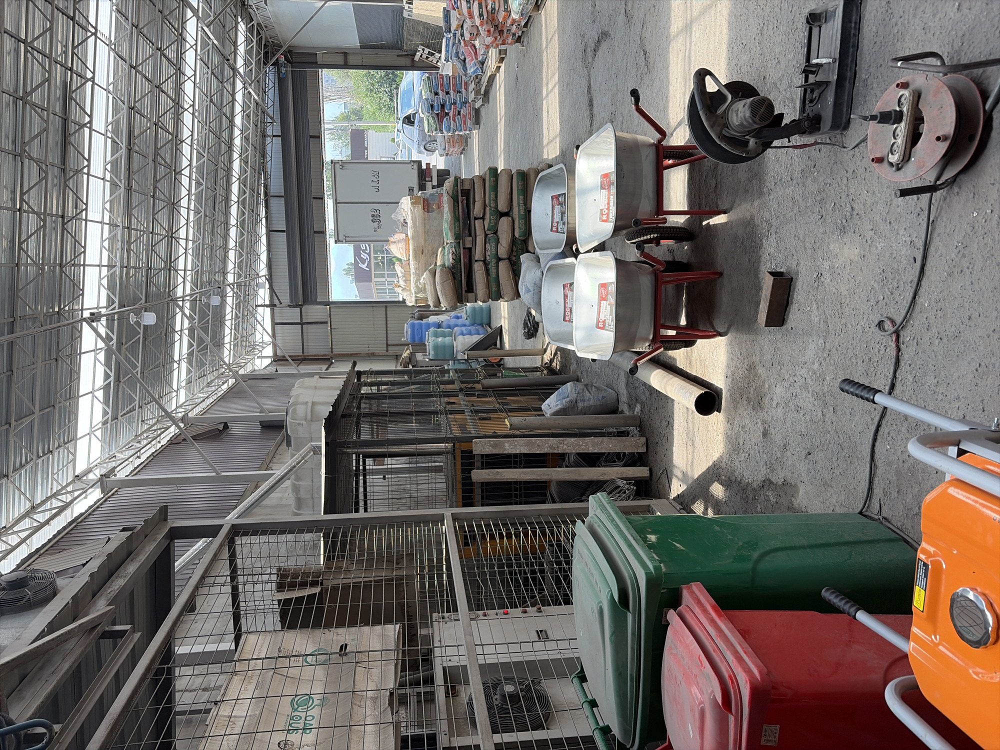 The image depicts the interior of a warehouse or storage facility in Novopokrovka, Kyrgyzstan. The scene is cluttered with various construction materials and equipment. In the foreground, two wheelbarrows are positioned side-by-side, with red frames and silver basins. To the left of the wheelbarrows, there are two large trash bins, one red and one green. An orange power tool, possibly a generator or compactor, is visible in the lower left corner. Further into the warehouse, several pallets stacked with bags of cement or similar materials are arranged. A large truck is parked in the background, partially obscuring an open bay door. The warehouse has a high, metal-framed roof with translucent panels allowing natural light to filter in. The floor is concrete and appears dusty. The overall impression is of a functional, utilitarian space used for storing and distributing construction supplies.