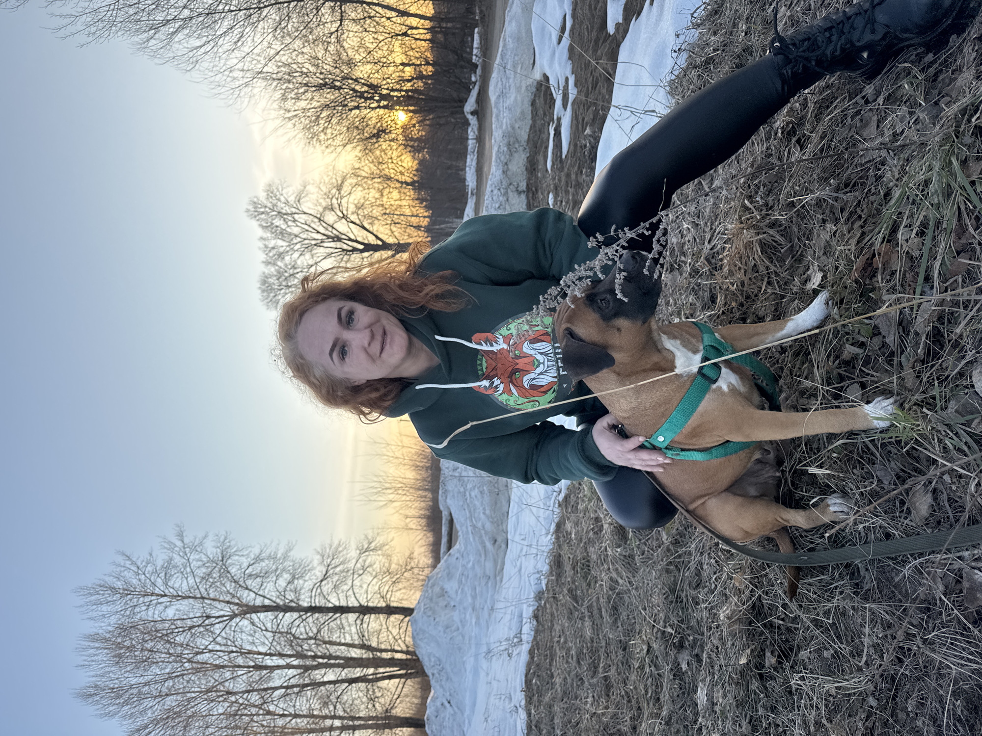 A red-haired woman in a green hoodie is squatting on a patch of dry grass and dead leaves next to a boxer dog with white markings on its paws and muzzle. The dog is wearing a green harness and leash. The woman is smiling and has her arm around the dog. The background shows a field with patches of snow, bare trees, and a soft sunset or sunrise. The scene is likely in a park or field in the city of Chaikovsky, Russia.