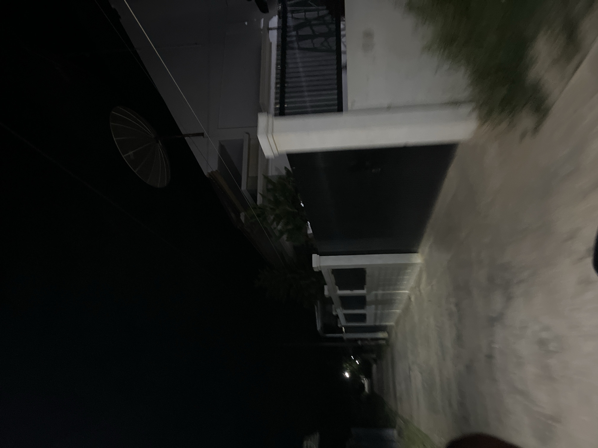 This is a nighttime, vertical shot of an outdoor scene in Menanggini, Indonesia. The image shows a residential area with a modern, multi-story building on the right side. The building has white accents and dark-colored sections, and there are horizontal blinds on some of the windows. A satellite dish is visible on a black wall to the left. The ground appears to be a paved walkway or driveway, wet from recent rain, reflecting the faint artificial lights from the building and street lamps. Trees and foliage are also visible, adding to the residential feel. The overall mood is quiet and serene, with the darkness suggesting late evening or early morning. There is no visible text in the image.