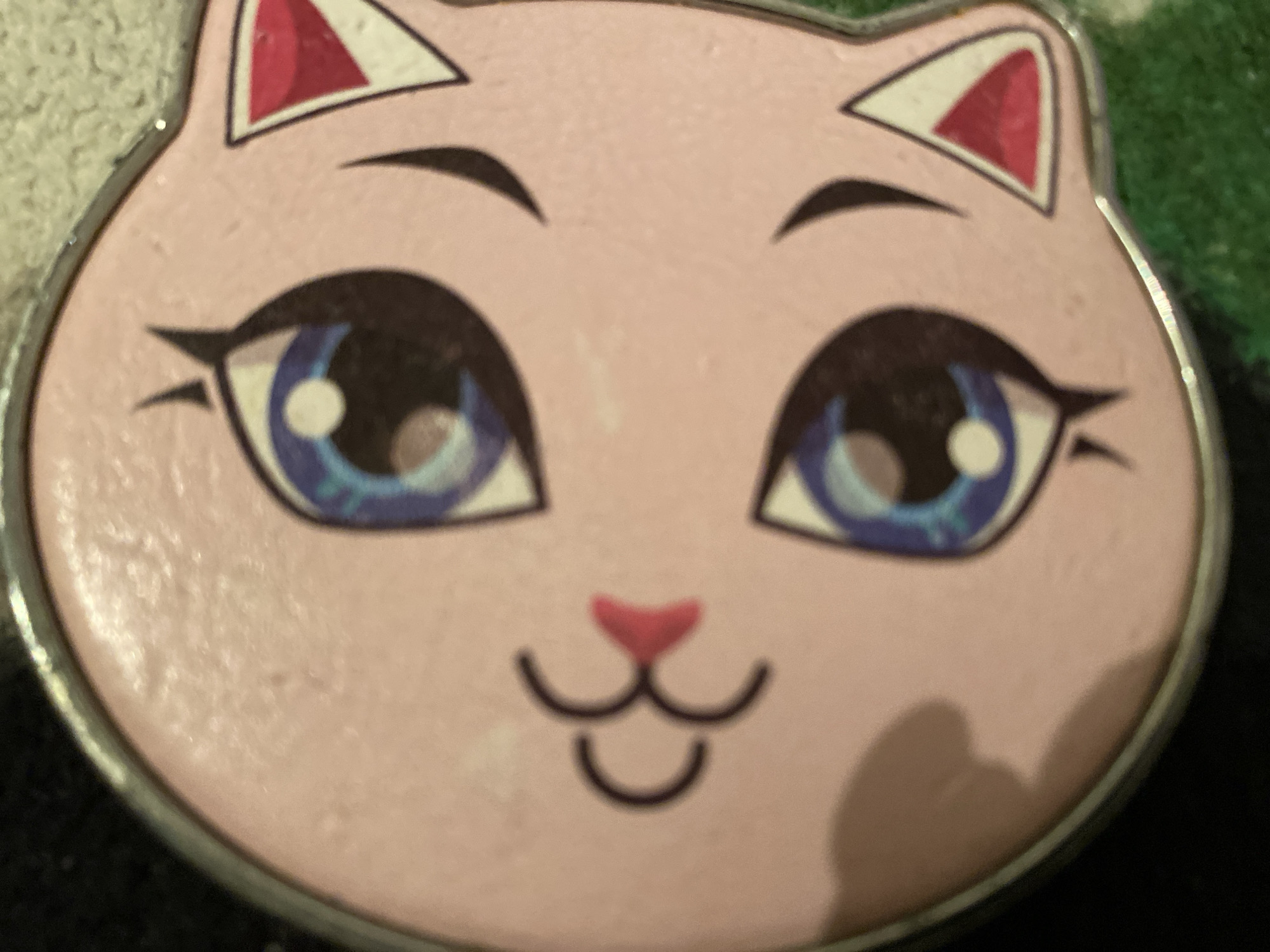 The image shows a close-up of a round, pink object with a cartoon cat face design. The cat has large blue eyes with white pupils, a pink nose, and black lines for a mouth and whiskers. There are also triangular pink and white shapes inside the cat's ears. The object appears to be made of plastic or a similar material, and has a metallic rim. A blurred green and white textured surface is visible in the background, and a dark background is also present to the right. The image is slightly out of focus and tilted. There is no visible indication of the city of Maloarslangulovo, Russia, or any specific location.